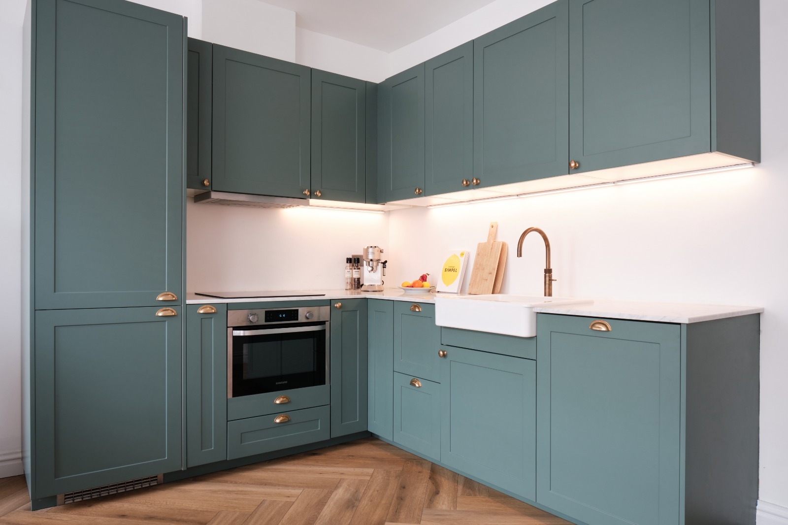 Zielona zabudowa kuchenna z frezowanymi frontami, złotymi uchwytami, białym zlewem farmerskim i blatem z marmuru, oświetlona listwą LED podszafkową.
