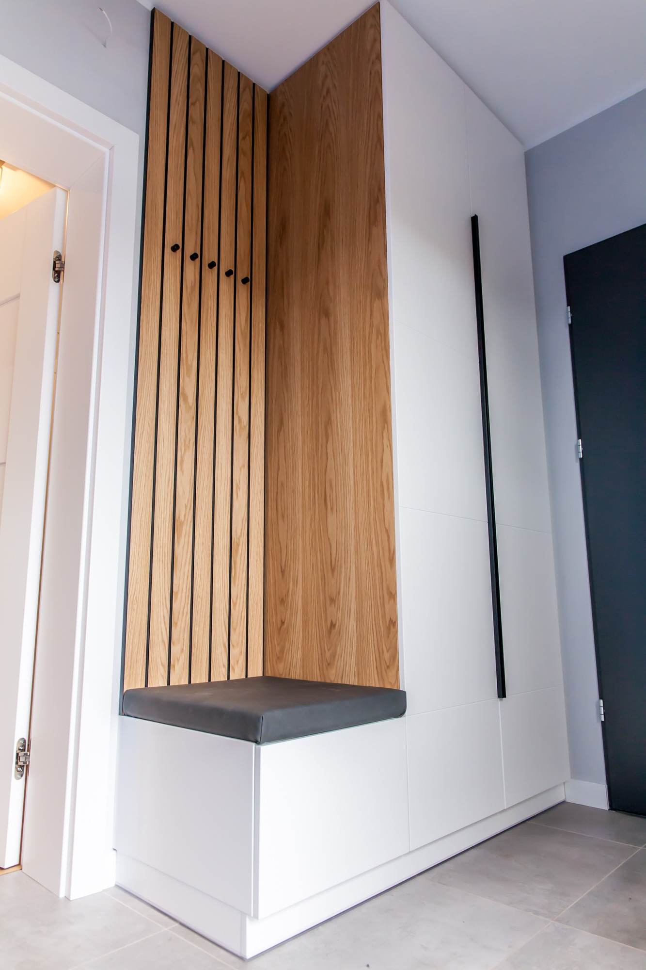 Biała szafa wnękowa z drewnianym panelem i siedziskiem w przedpokoju. Minimalistyczny design z czarnymi akcentami i wieszakami.