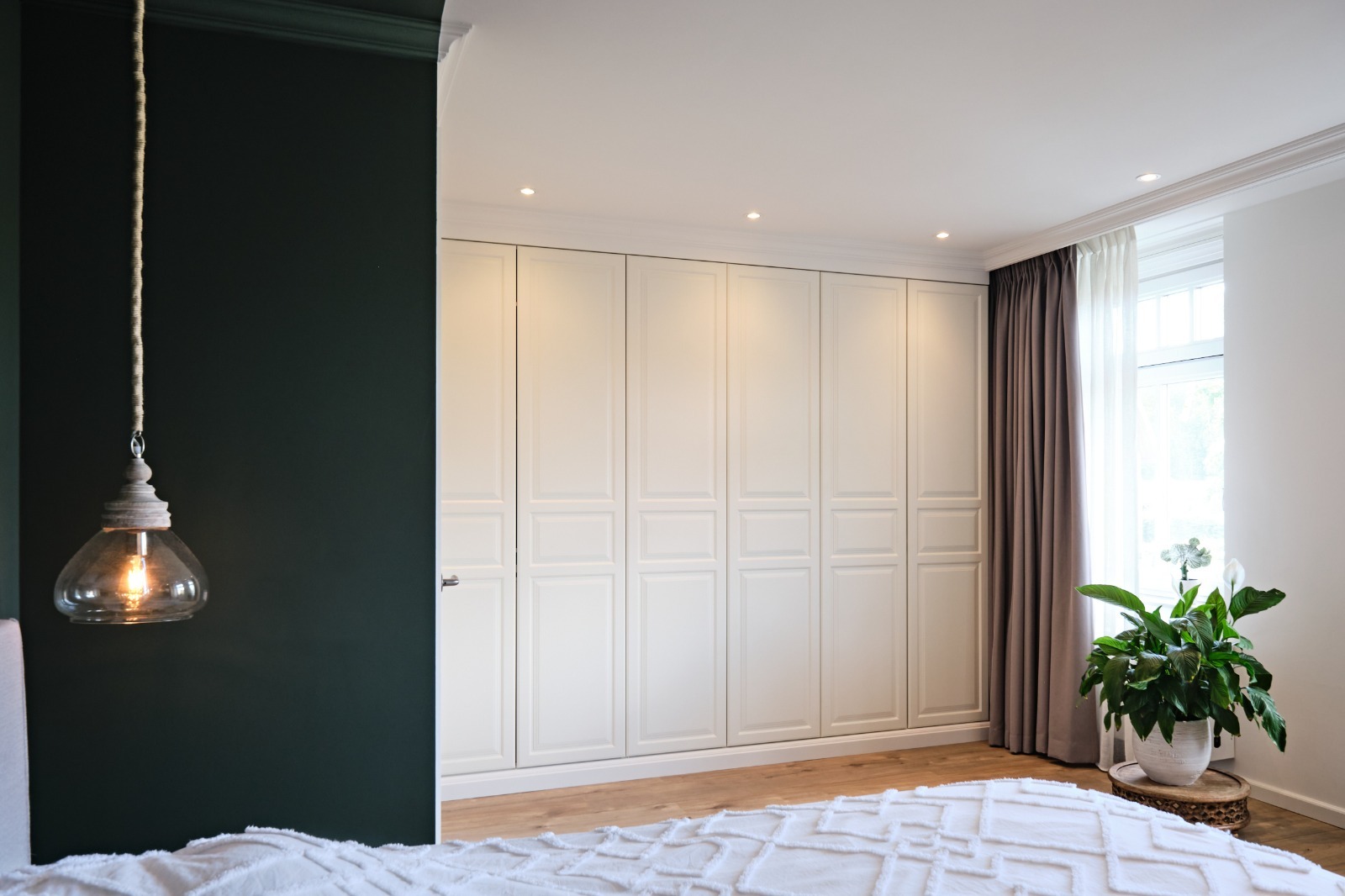 Biała szafa wnękowa z frezowanymi drzwiami, oświetlona punktowo, w sypialni z ciemnozieloną ścianą i lampą wiszącą. Minimalistyczny design, jasne drewno na podłodze.