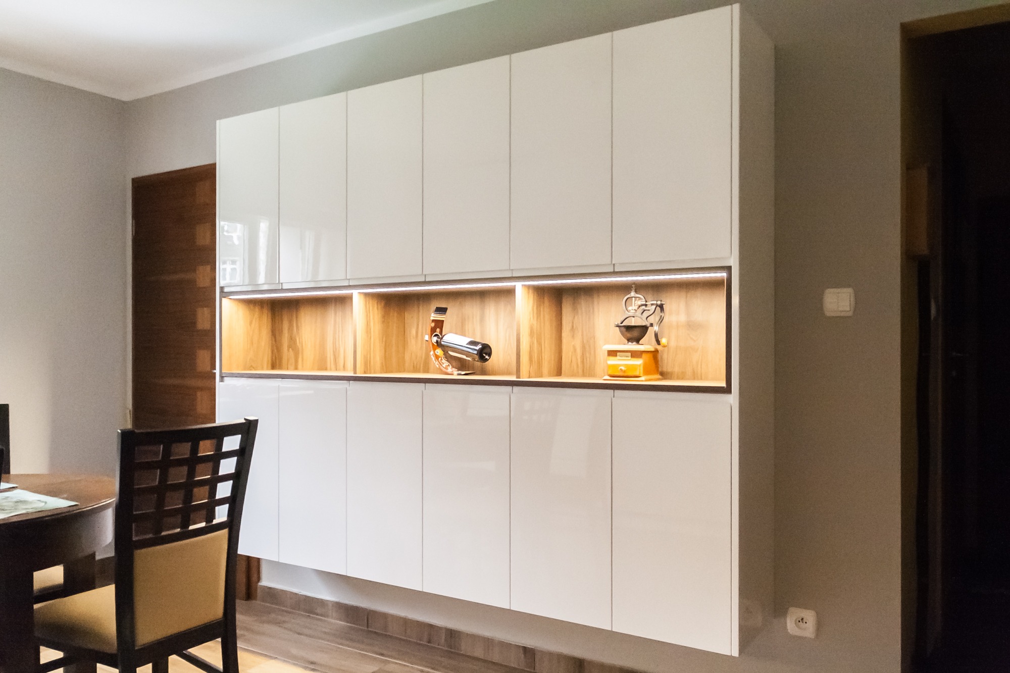 Biała szafa wisząca z oświetlonymi wnękami, w których stoją dekoracje: młynek do kawy i stojak na wino. Nowoczesny design, widok z jadalni.
