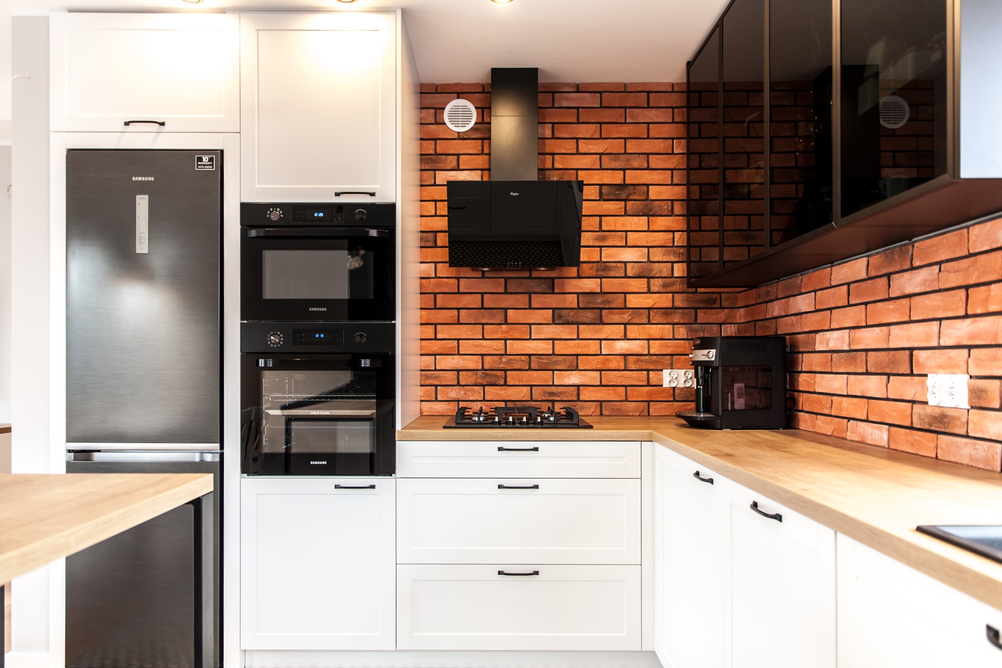 Nowoczesna kuchnia z białymi szafkami, drewnianym blatem i ścianą z czerwonej cegły. Widoczny sprzęt AGD: lodówka, piekarnik, płyta gazowa i ekspres do kawy.