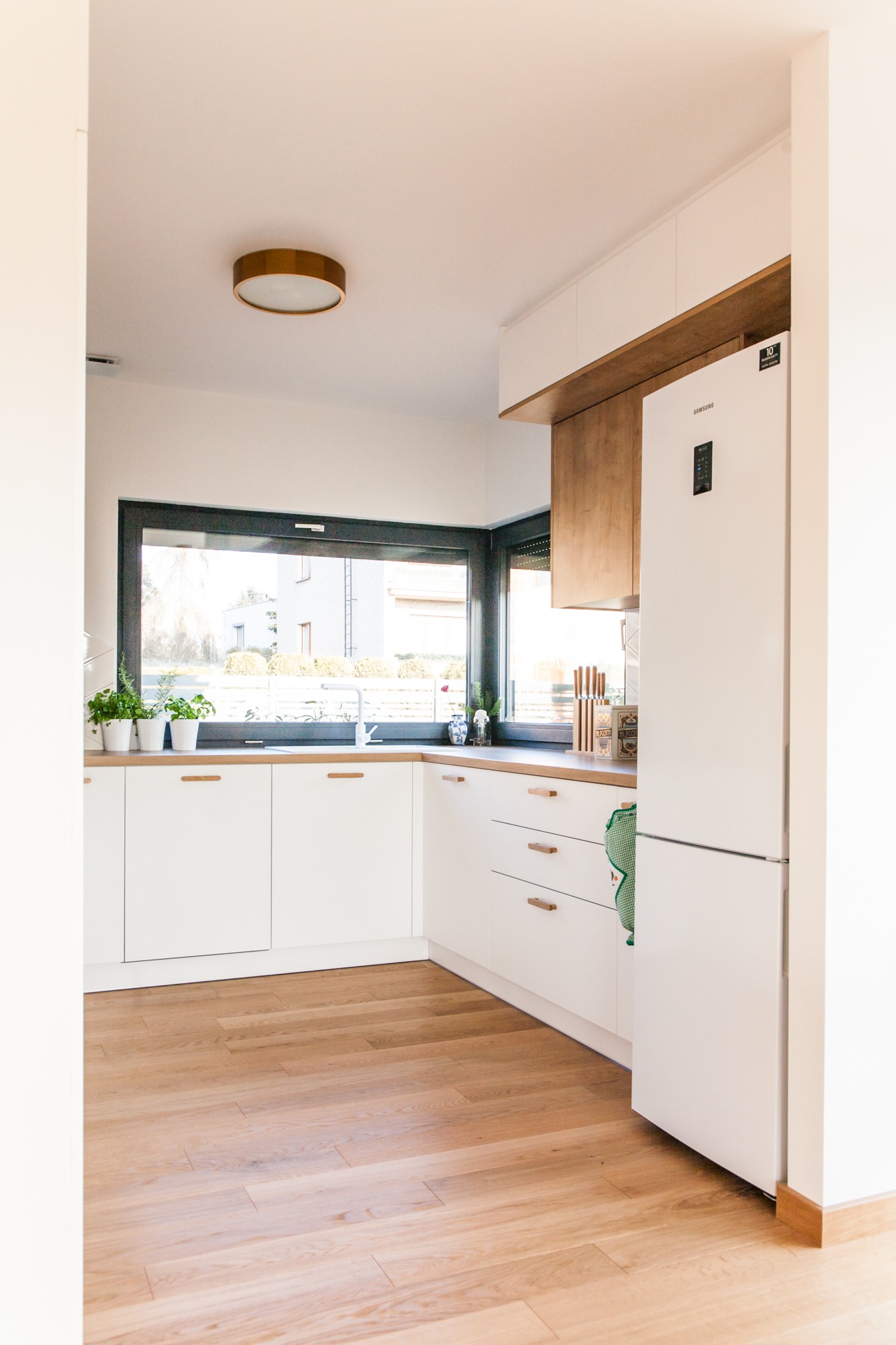 Jasna kuchnia z białymi szafkami i drewnianymi blatami, okno z widokiem na zieleń, nowoczesna lampa sufitowa i lodówka Samsung. Minimalistyczny design.