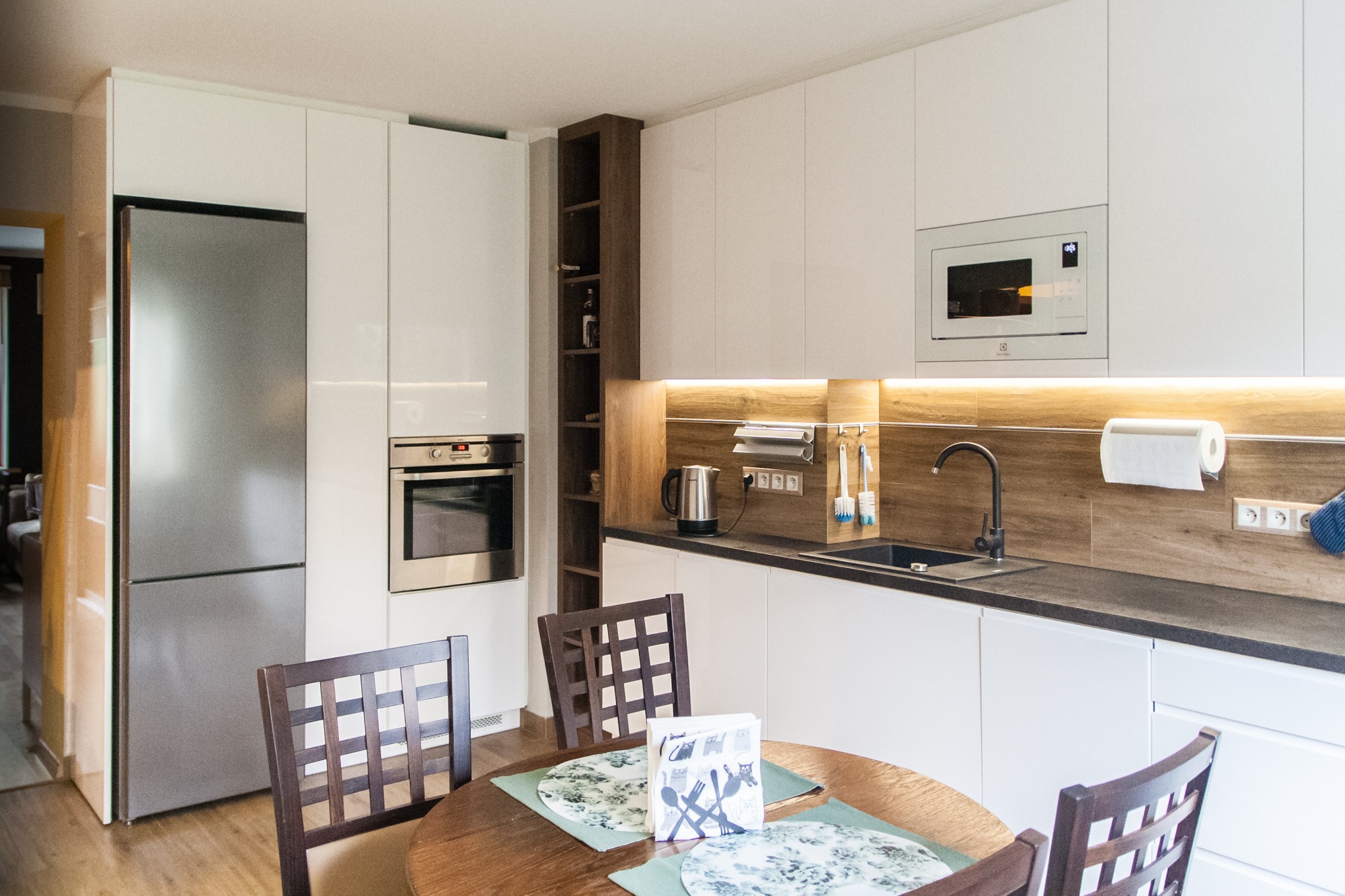 Nowoczesna kuchnia z białymi frontami, drewnianym blatem i oświetleniem LED. Na pierwszym planie okrągły stół z krzesłami. Widoczny sprzęt AGD: lodówka, piekarnik, mikrofalówka.