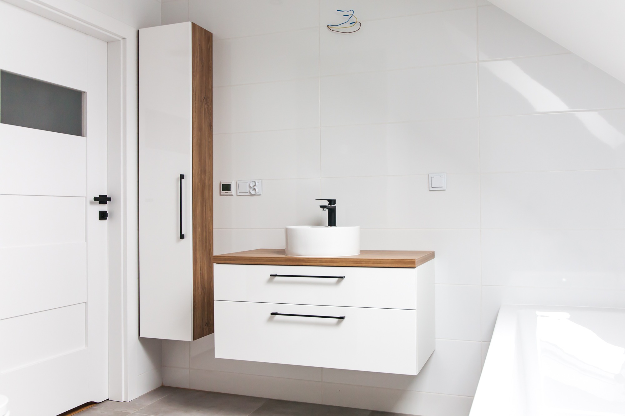 Nowoczesna łazienka z białą szafką pod umywalkę z drewnianym blatem i wysoką szafką obok białych drzwi z czarnymi detalami, na tle białych płytek.