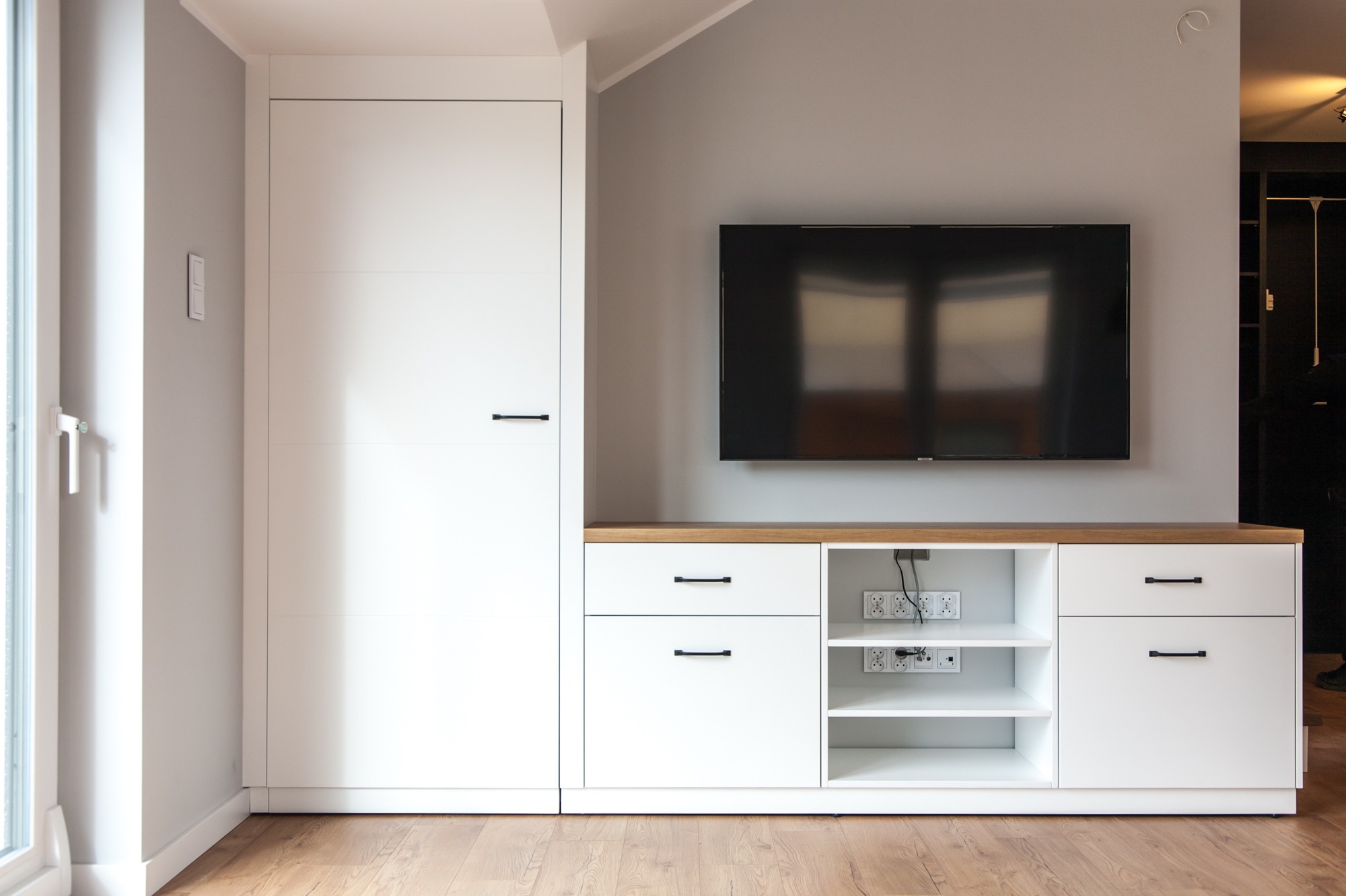Biała zabudowa meblowa z szafą i szafką RTV pod telewizor w salonie, Radomsko. Minimalistyczny design, drewniany blat, czarne uchwyty, widoczne gniazdka.