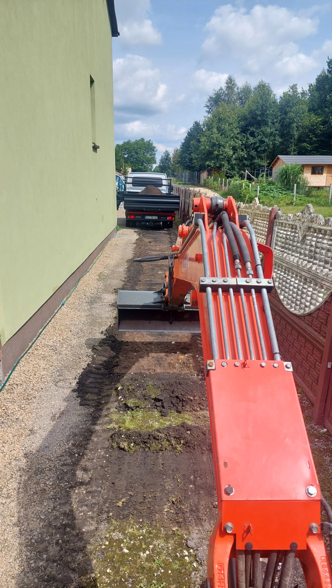 Pomarańczowa koparka podczas prac ziemnych przy budynku, w tle ciężarówka z ziemią, widok z kabiny operatora.