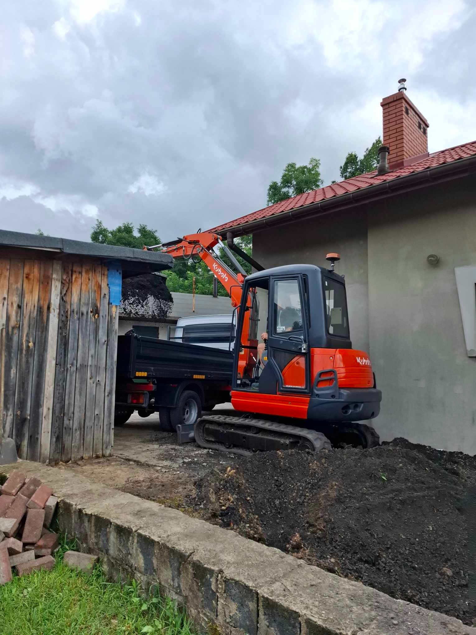 Pomarańczowa mini koparka Kubota ładuje ziemię do skrzyni wywrotki na tle szarego budynku i drewnianej szopy.