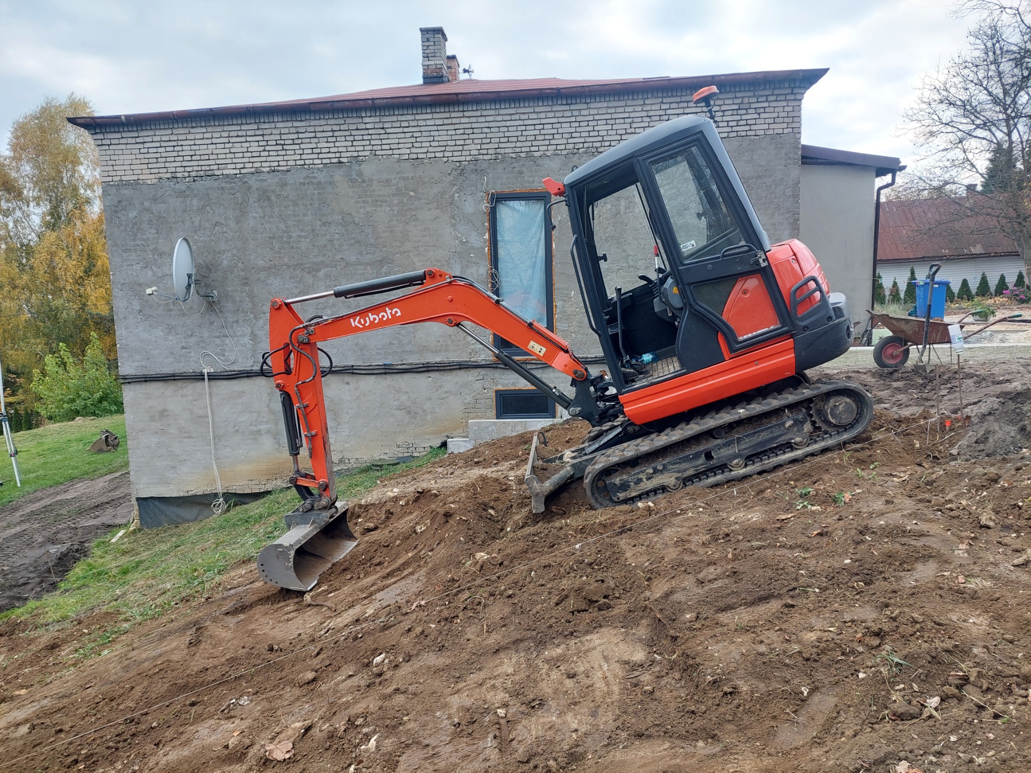 Pomarańczowa minikoparka Kubota na nierównym terenie z widocznym budynkiem w tle i otwartymi drzwiami kabiny.