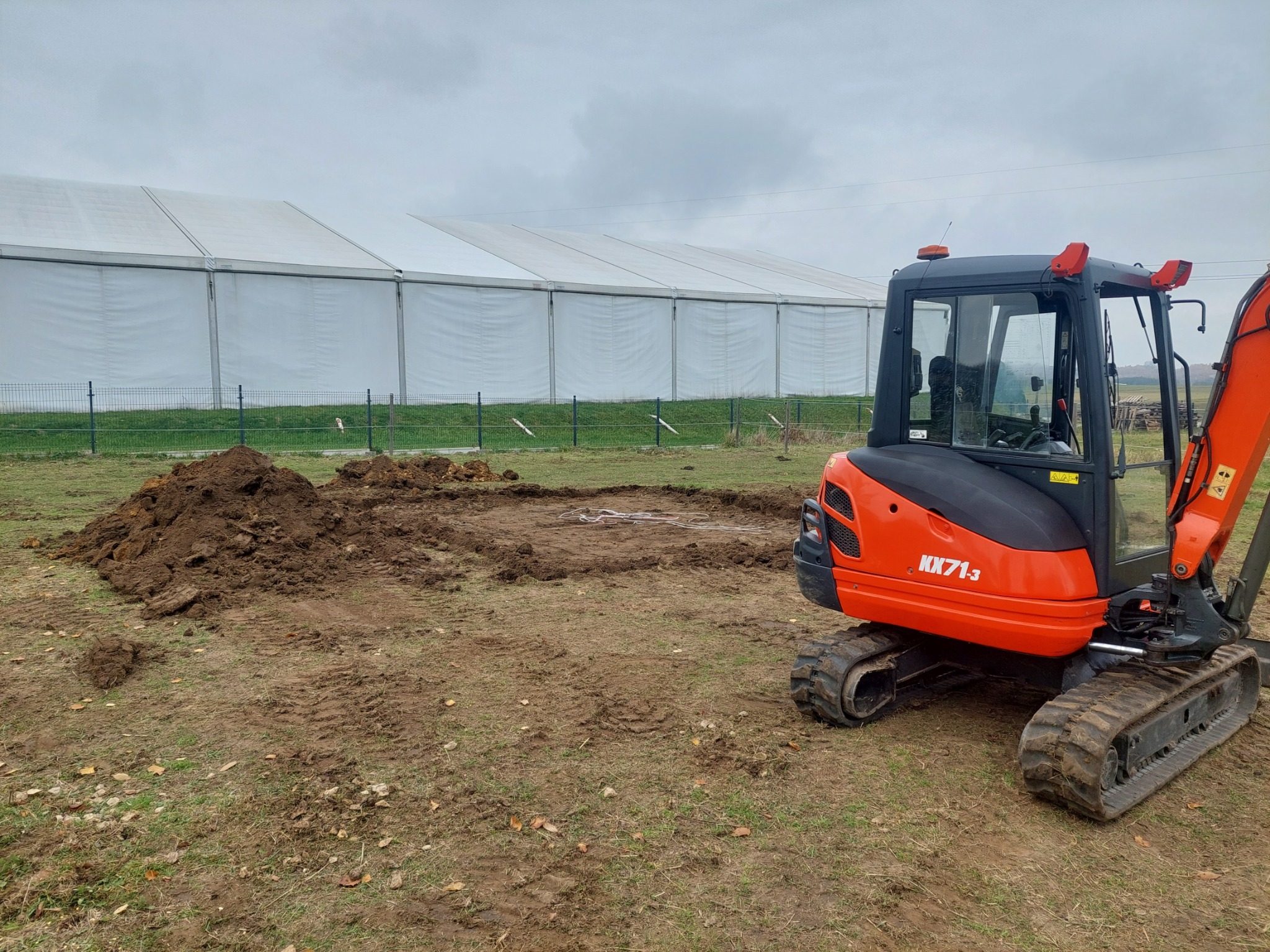 Pomarańczowa mini koparka Kubota KX71-3 na placu budowy, obok pryzma ziemi, w tle biały namiot halowy i metalowe ogrodzenie.