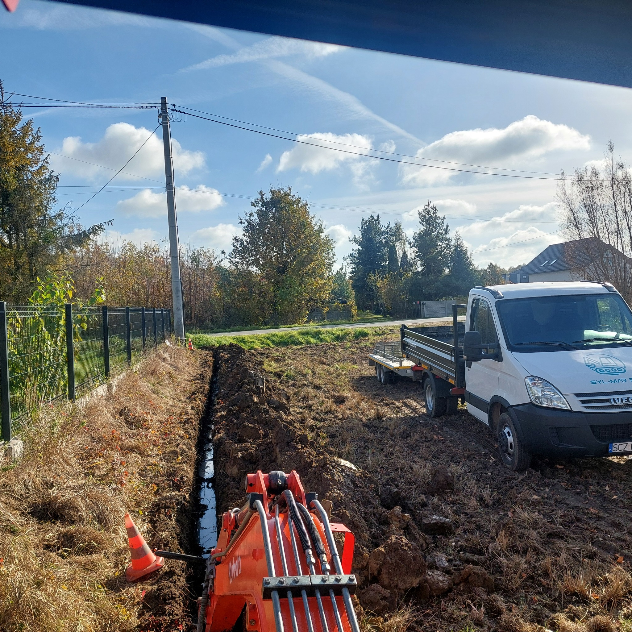 Pomarańczowa minikoparka przy wykopanym rowie wypełnionym wodą, obok biały samochód dostawczy z przyczepką na tle pola i błękitnego nieba z chmurami.