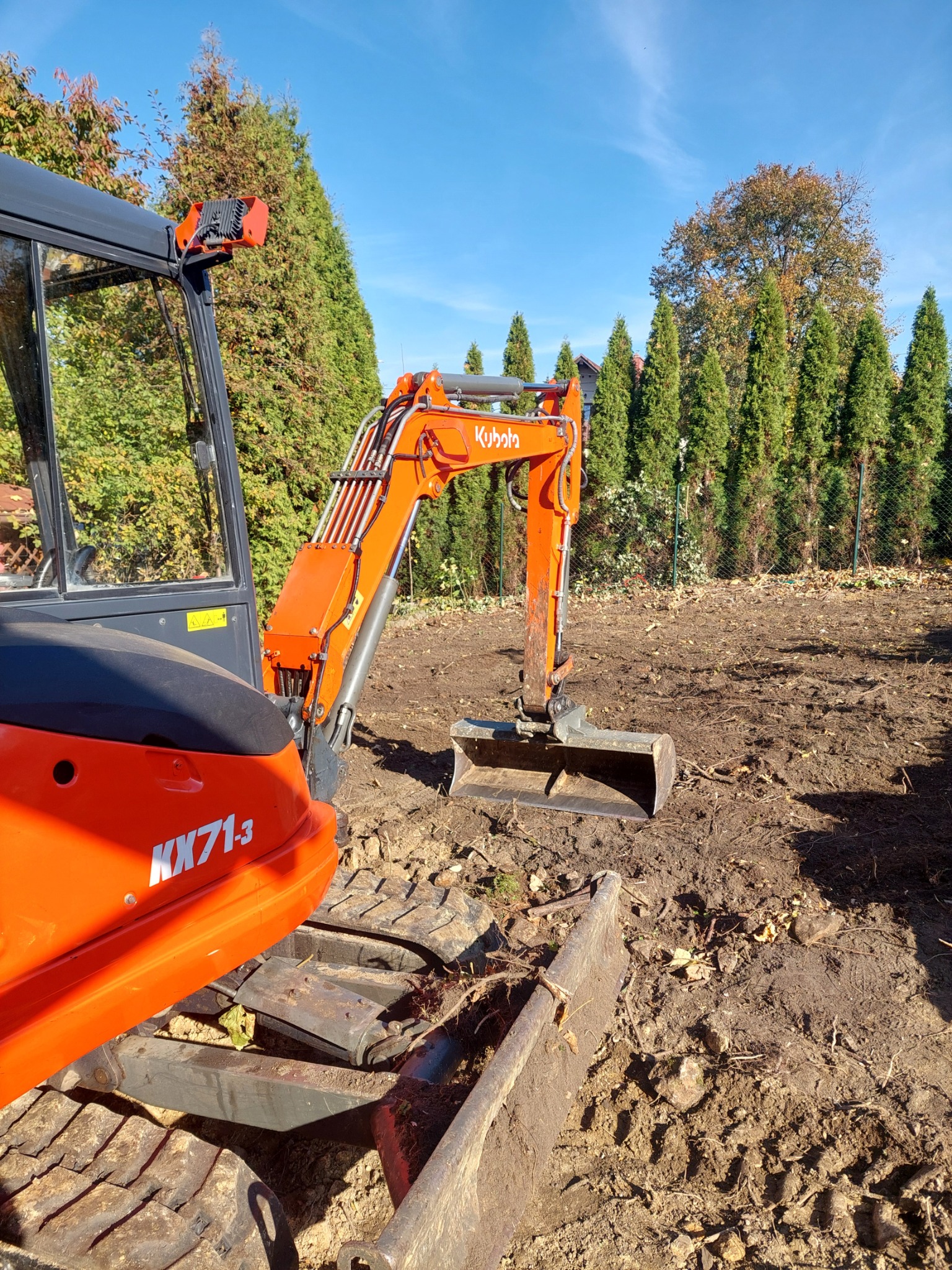 Pomarańczowa mini koparka Kubota KX71-3 na działce z przygotowanym terenem, widoczne ślady pracy i tło z zielonych drzew.