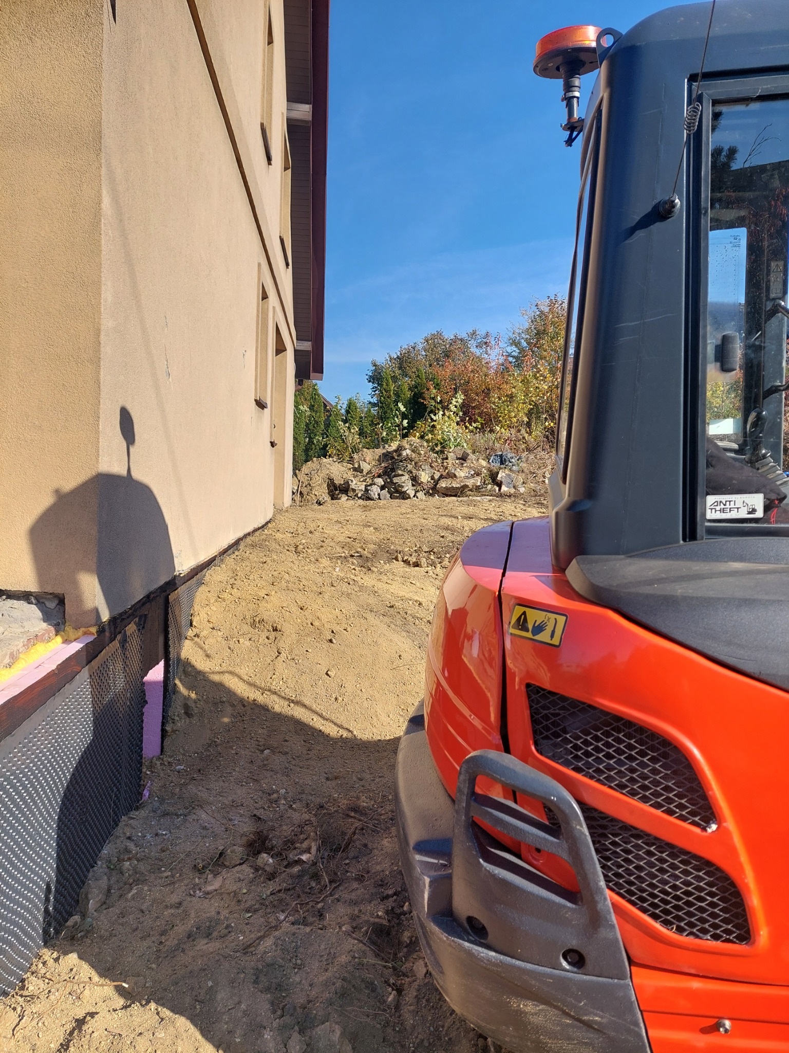Częściowo wykopana ziemia wzdłuż fundamentu budynku, widoczna folia kubełkowa i izolacja, obok stoi pomarańczowa minikoparka.