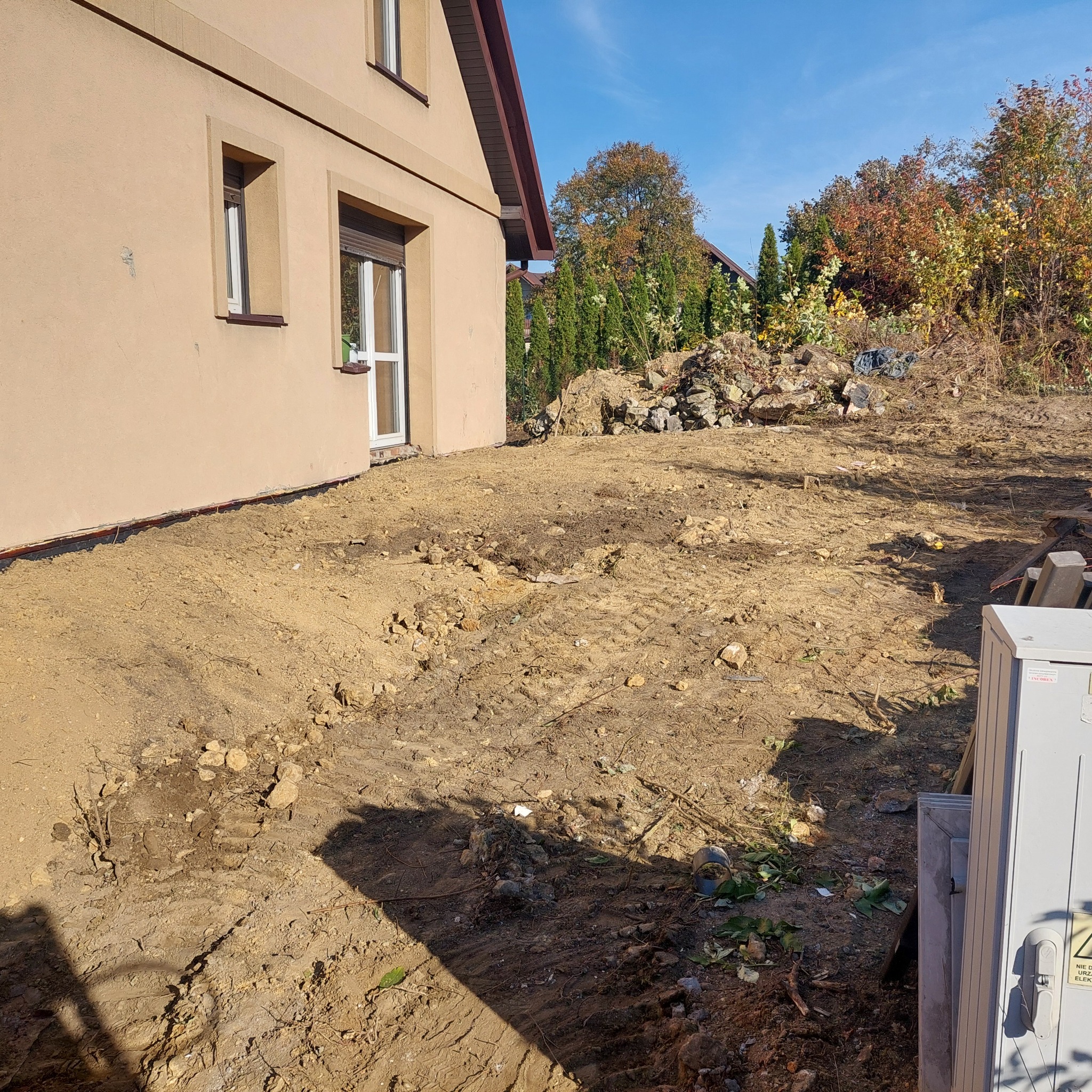 Przygotowany teren pod budowę, widoczne ślady opon i resztki roślinności, budynek w tle.