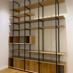 Regał drewniany z elementami metalowymi w stylu industrialnym, z zabudowanymi szafkami na dole i otwartymi półkami, oświetlenie sufitowe punktowe.