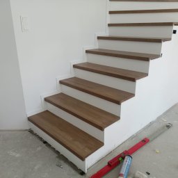 Mieszko Prajsnar PrajsIdea - Nowoczesne schody z drewnianymi stopniami i białymi podstopnicami, widoczne narzędzia montażowe na surowej podłodze, świeżo po instalacji.