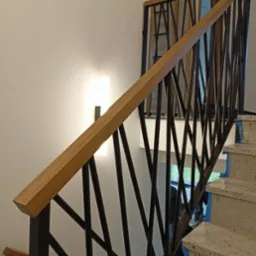 Nowoczesne schody z drewnianą poręczą i metalową balustradą o geometrycznym wzorze, oświetlone lampą ścienną, prowadzące na wyższe piętro. Detal wykończenia.