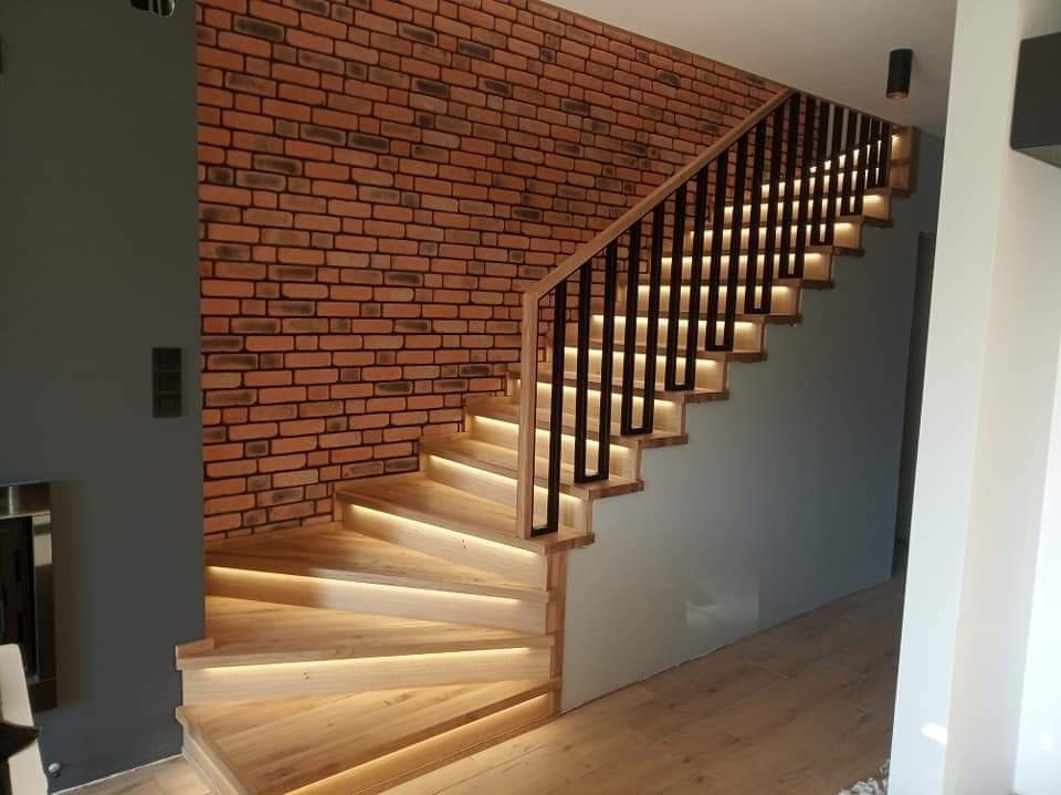 Kątowe ujęcie drewnianych schodów z podświetleniem LED na każdym stopniu, z czarną balustradą i ścianą z cegły w tle.