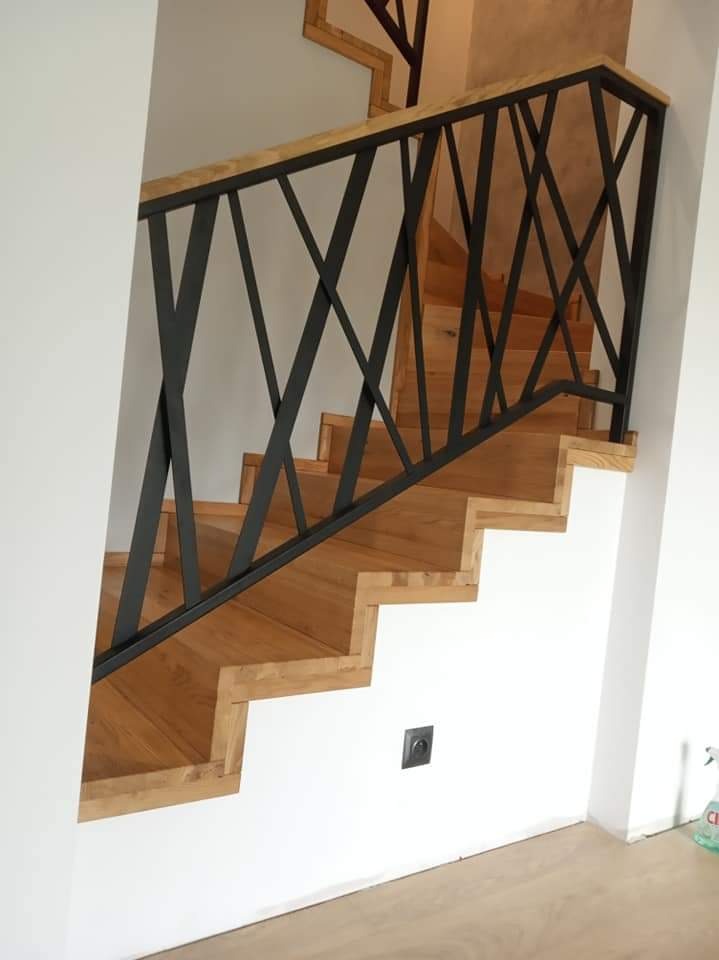 Nowoczesne schody z drewna z czarną metalową balustradą o geometrycznym wzorze, widoczne z dołu, z jasną podłogą i białymi ścianami, z gniazdkiem elektrycznym na ścianie.