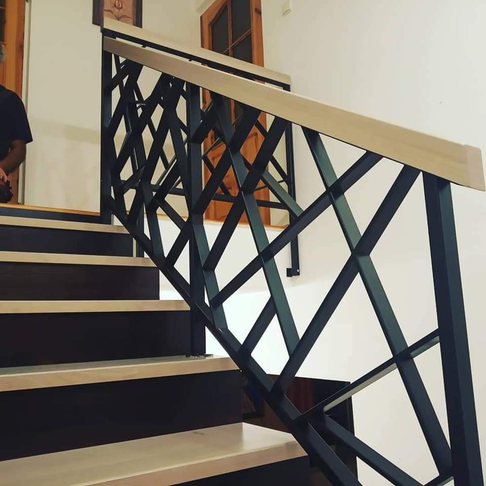 Nowoczesne schody z metalową balustradą o geometrycznym wzorze, drewniany pochwyt, widoczne stopnie w odcieniach brązu i beżu.