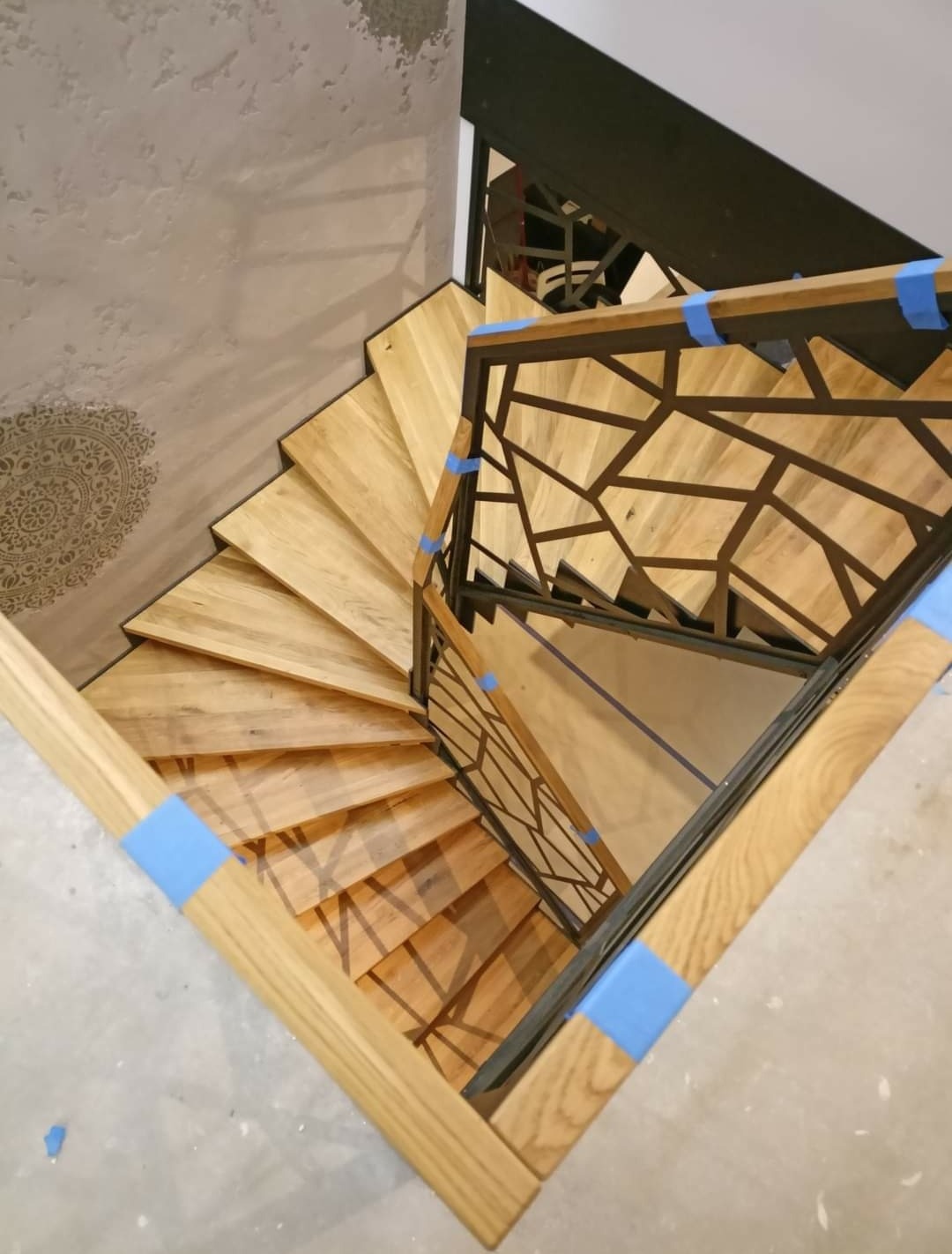 Wewnętrzne, drewniane schody spiralne z metalową balustradą o geometrycznym wzorze, zabezpieczone niebieską taśmą malarską, widok z góry na surowe wykończenie podłogi.