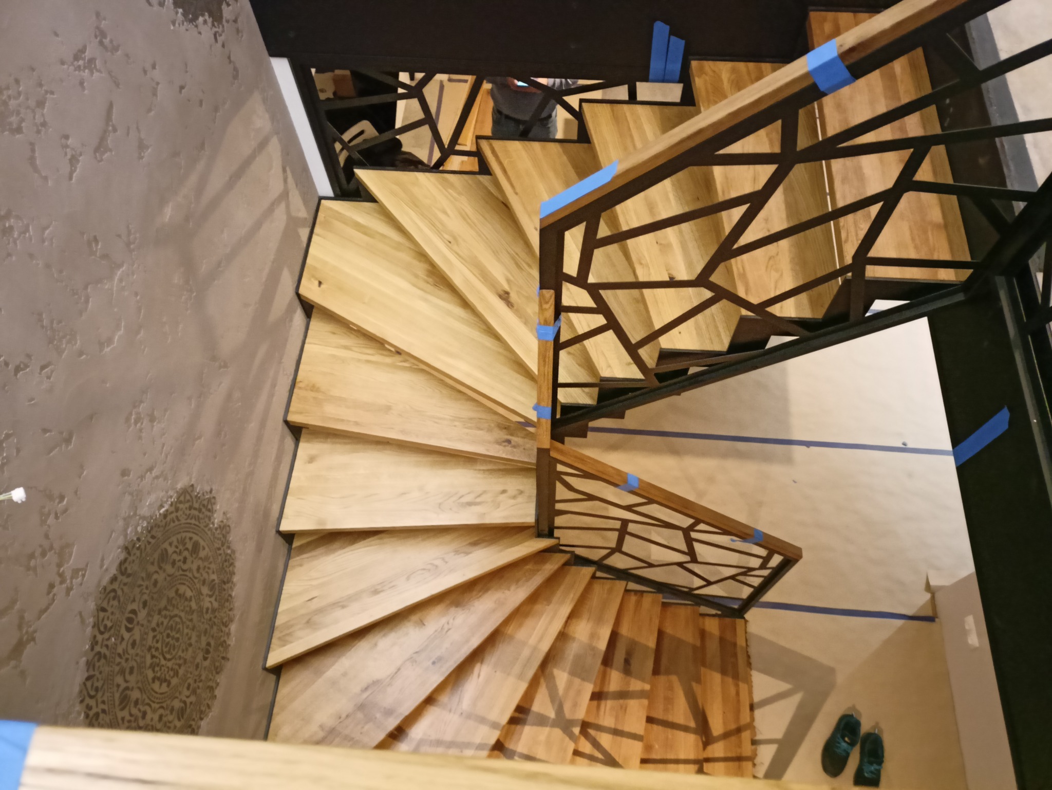 Spiralne schody z jasnego drewna z metalową balustradą o geometrycznym wzorze, zabezpieczone niebieską taśmą malarską, widok z góry.