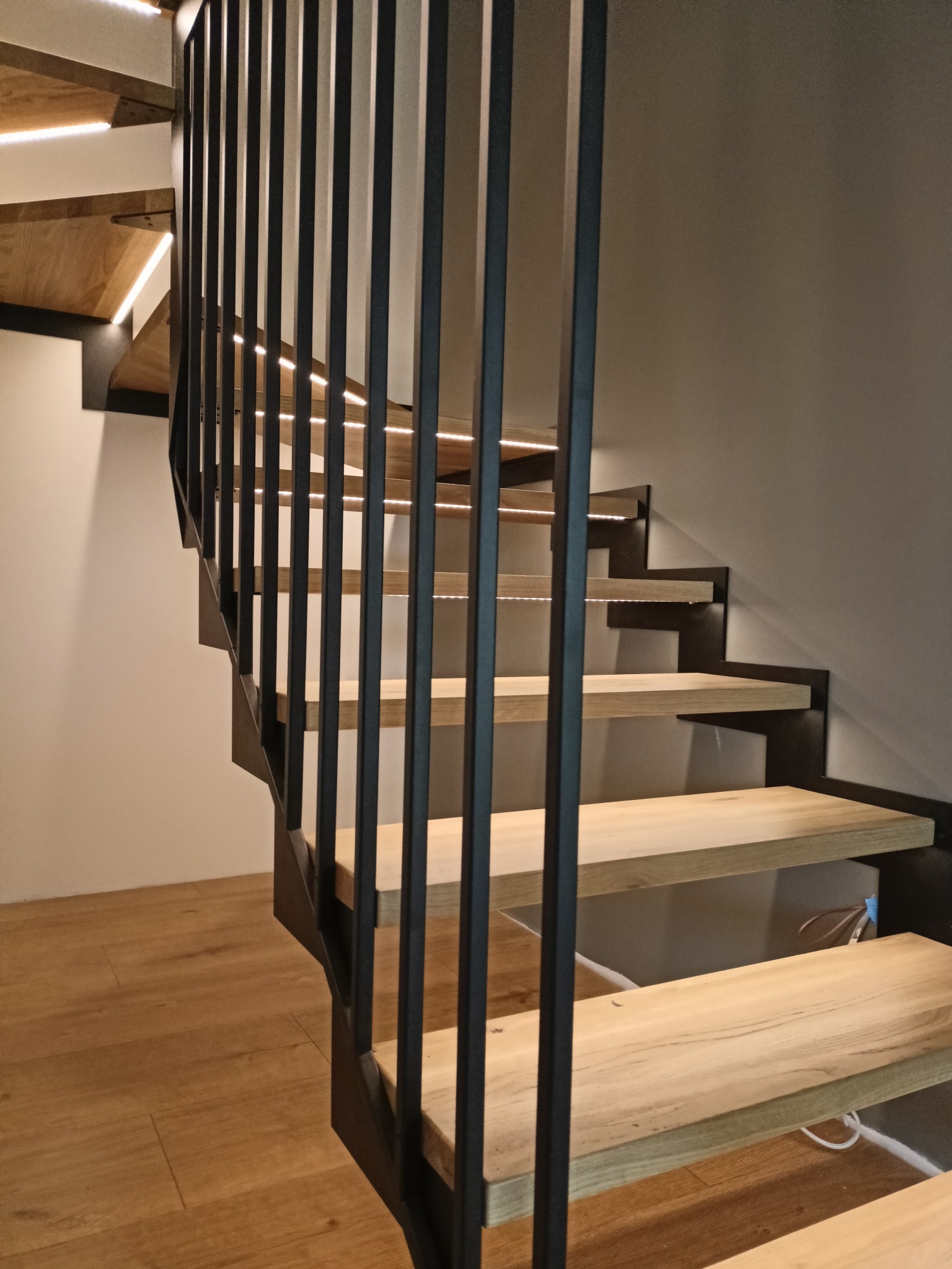 Nowoczesne schody z drewnianymi stopniami i metalową balustradą z pionowych prętów, podświetlane od spodu, widoczne od dołu