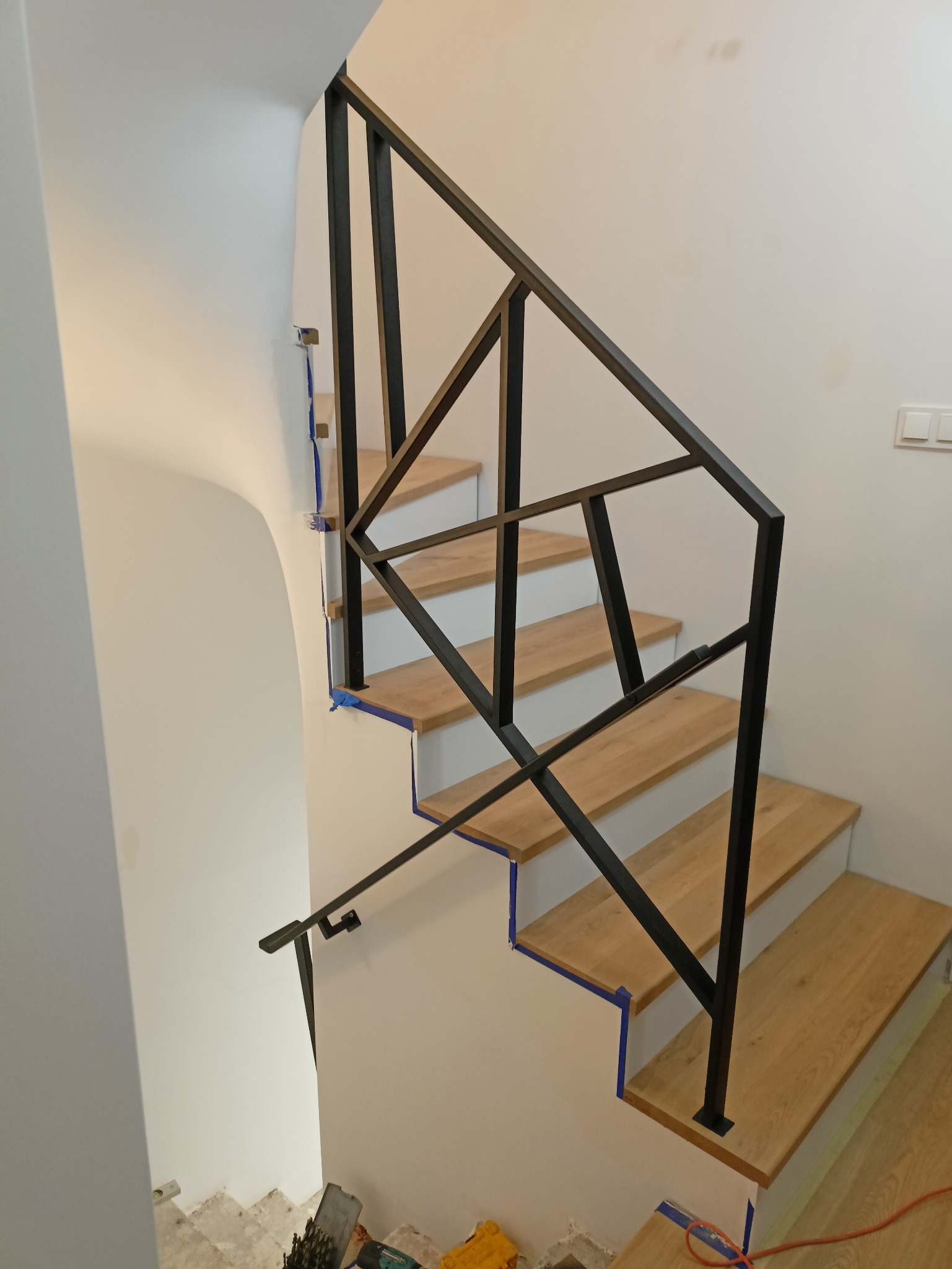 Montaż schodów z drewnianymi stopniami i czarną, geometryczną balustradą metalową, zabezpieczone niebieską taśmą malarską, widoczne narzędzia i materiały budowlane u dołu.
