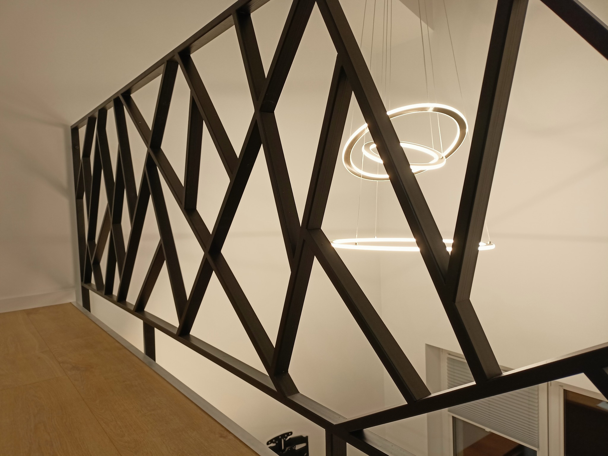Nowoczesna balustrada metalowa z geometrycznym wzorem na schodach, oświetlona wiszącą lampą pierścieniową LED.