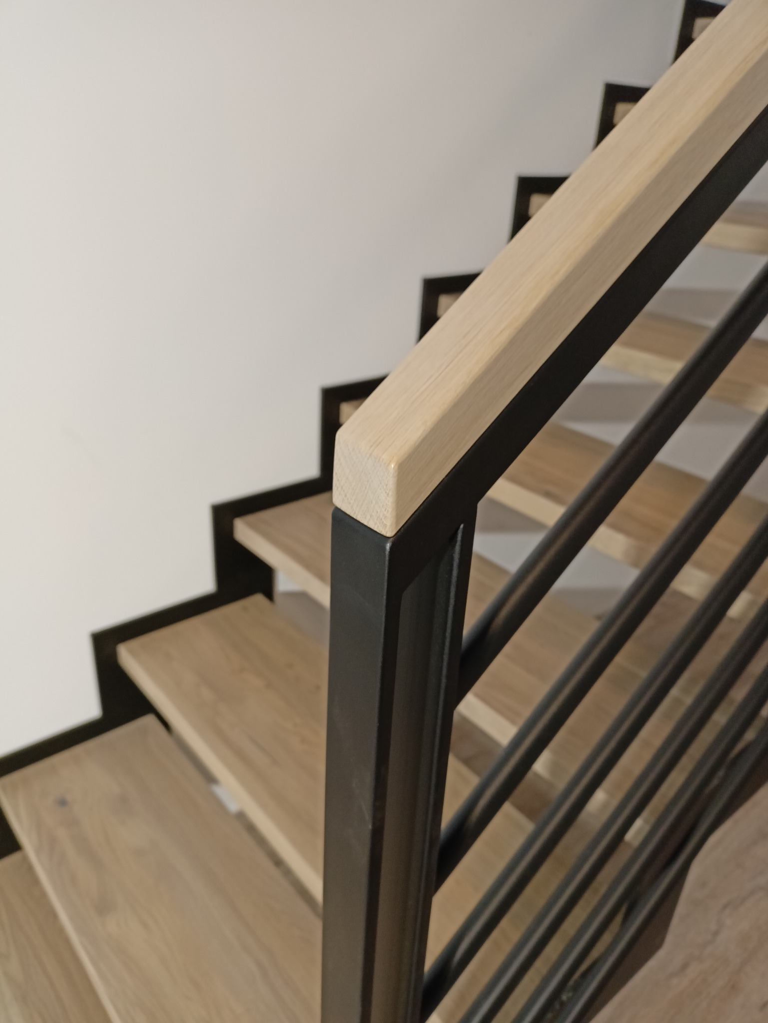 Nowoczesne schody z drewnianymi stopniami i czarną metalową balustradą z jasnym drewnianym poręczą, widok z góry na fragment konstrukcji.