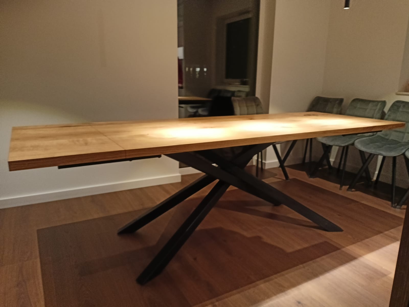 Rozkładany stół z blatem z jasnego drewna i czarną, metalową podstawą w kształcie litery X, w tle krzesła tapicerowane welurem w odcieniach szarości i zieleni.