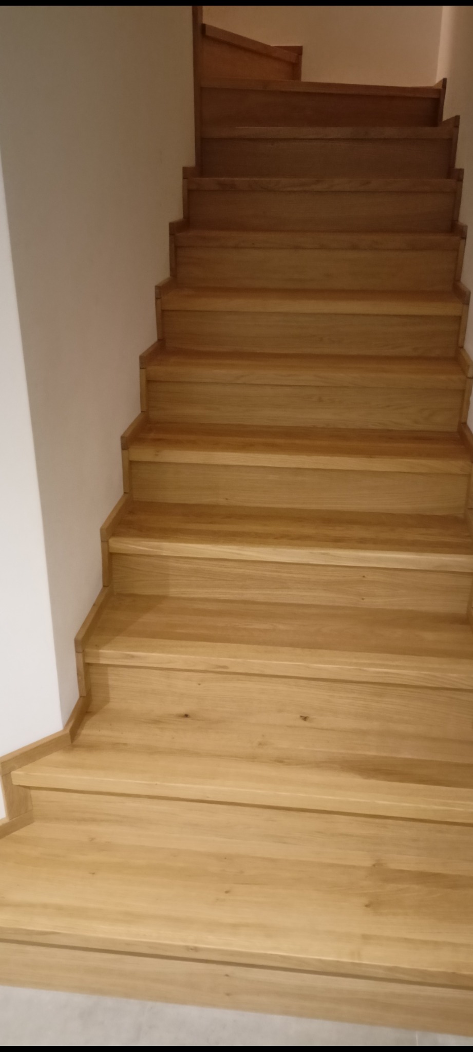 Jasne, drewniane schody z zaokrąglonymi krawędziami stopni, prowadzące w górę wzdłuż białej ściany.