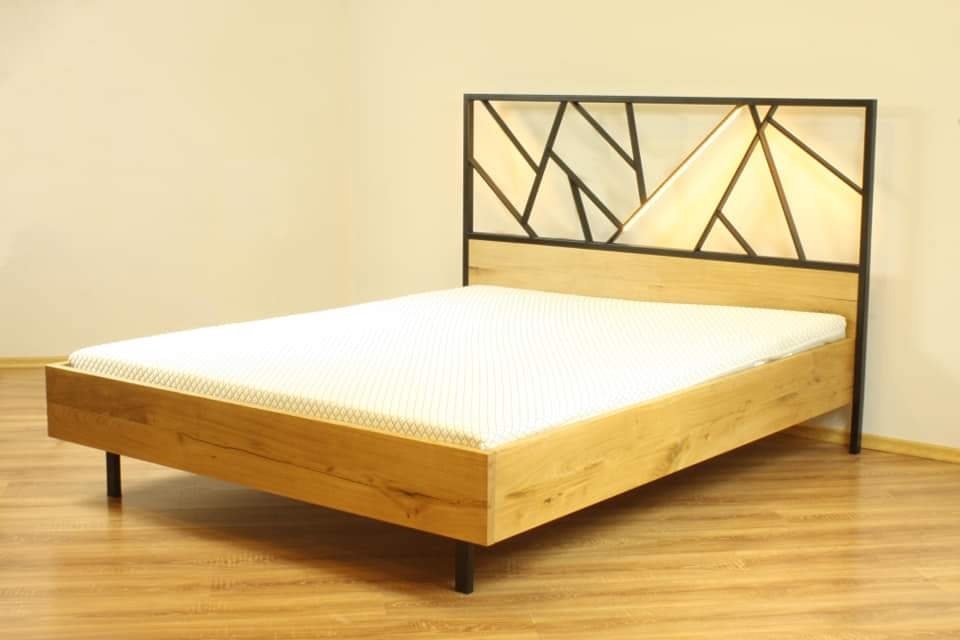 Drewniane łóżko z metalowym zagłówkiem o geometrycznym wzorze, oświetlonym od tyłu, na drewnianej podłodze.