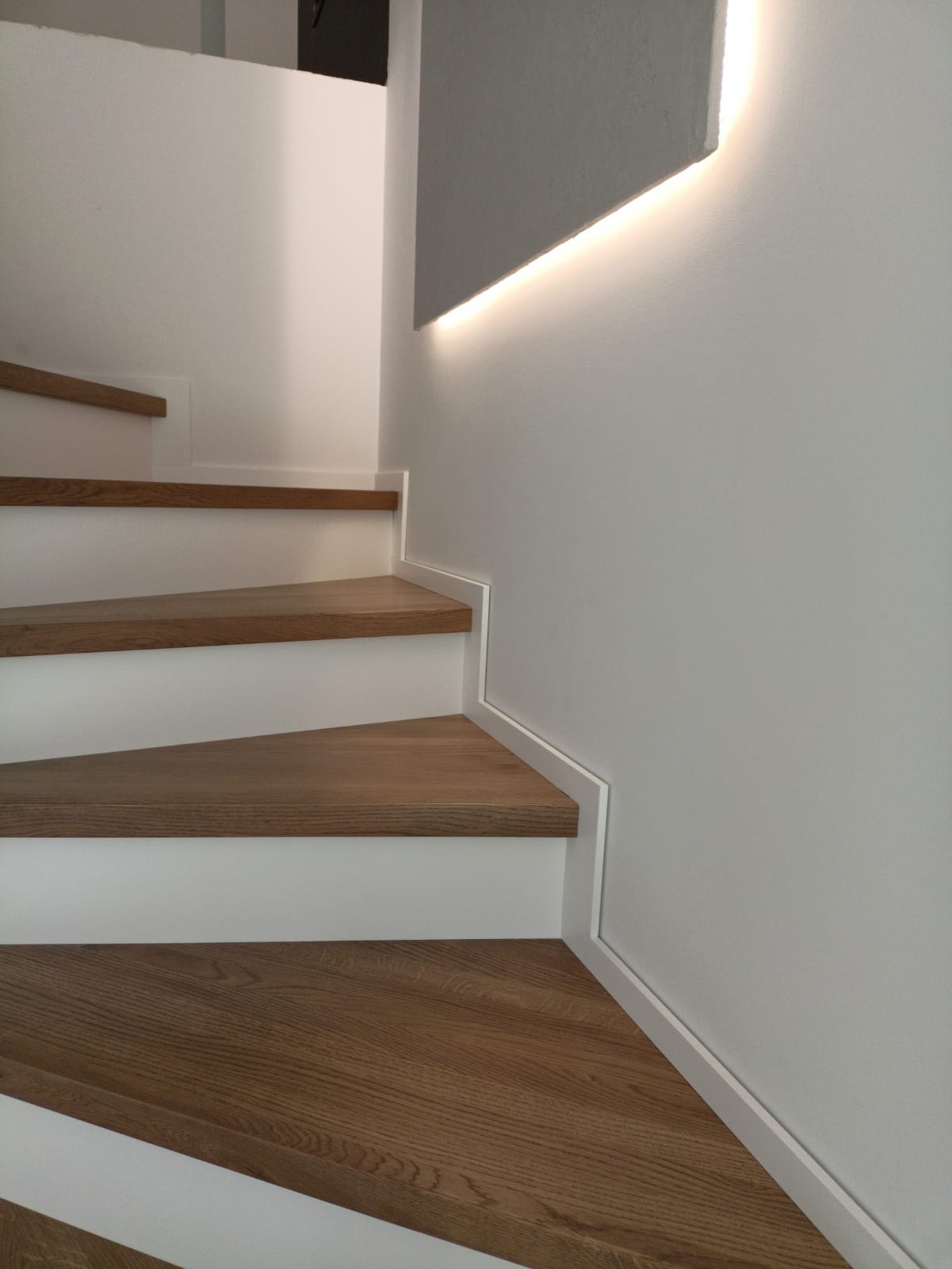 Nowoczesne schody z drewnianymi stopniami i białymi podstopnicami, oświetlenie LED na ścianie, minimalistyczny design, jasne wnętrze.