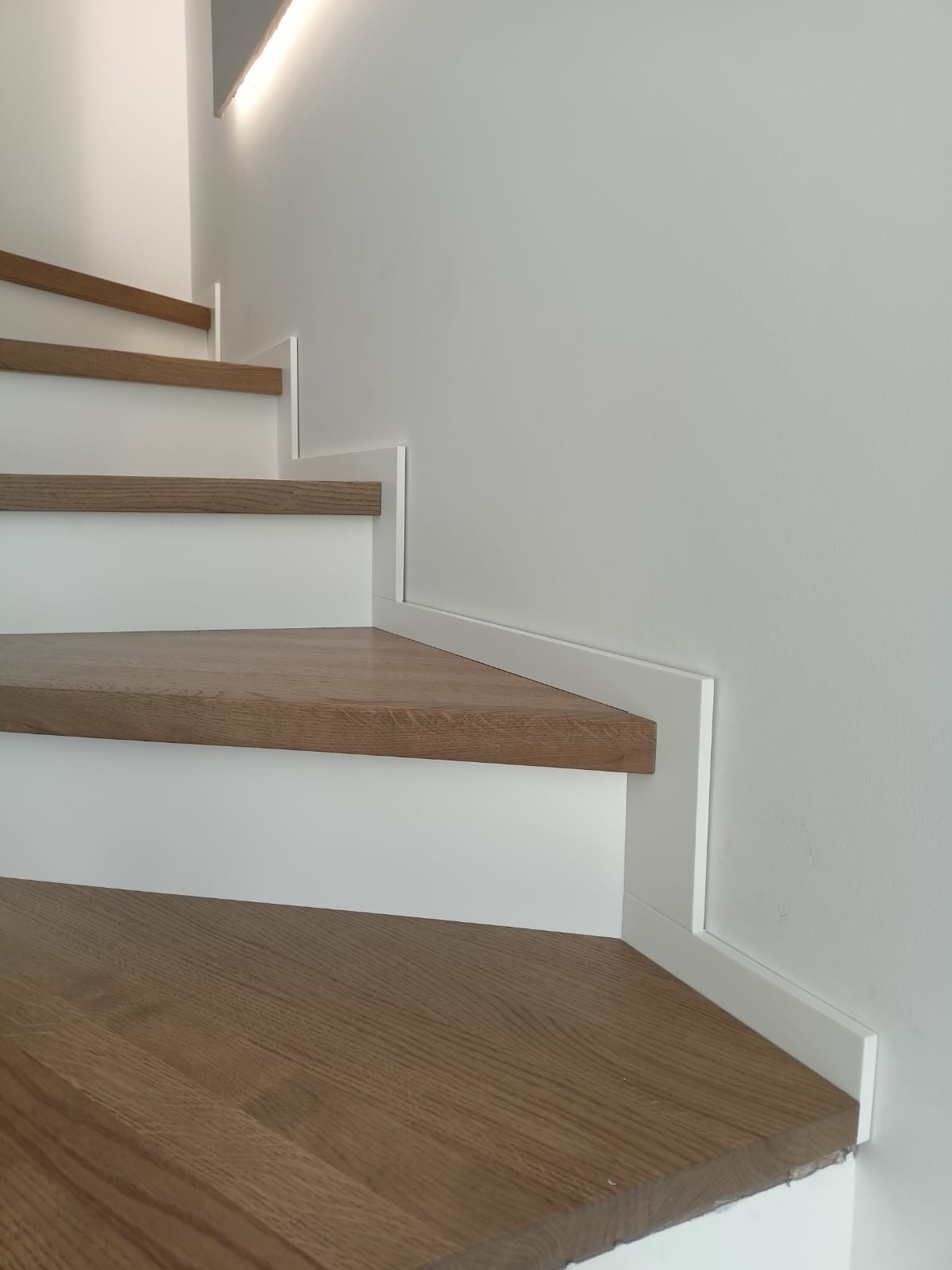 Minimalistyczne schody: drewniane stopnie o ciepłym odcieniu, białe podstopnice i listwy przypodłogowe, oświetlenie LED na górze. Nowoczesny design.