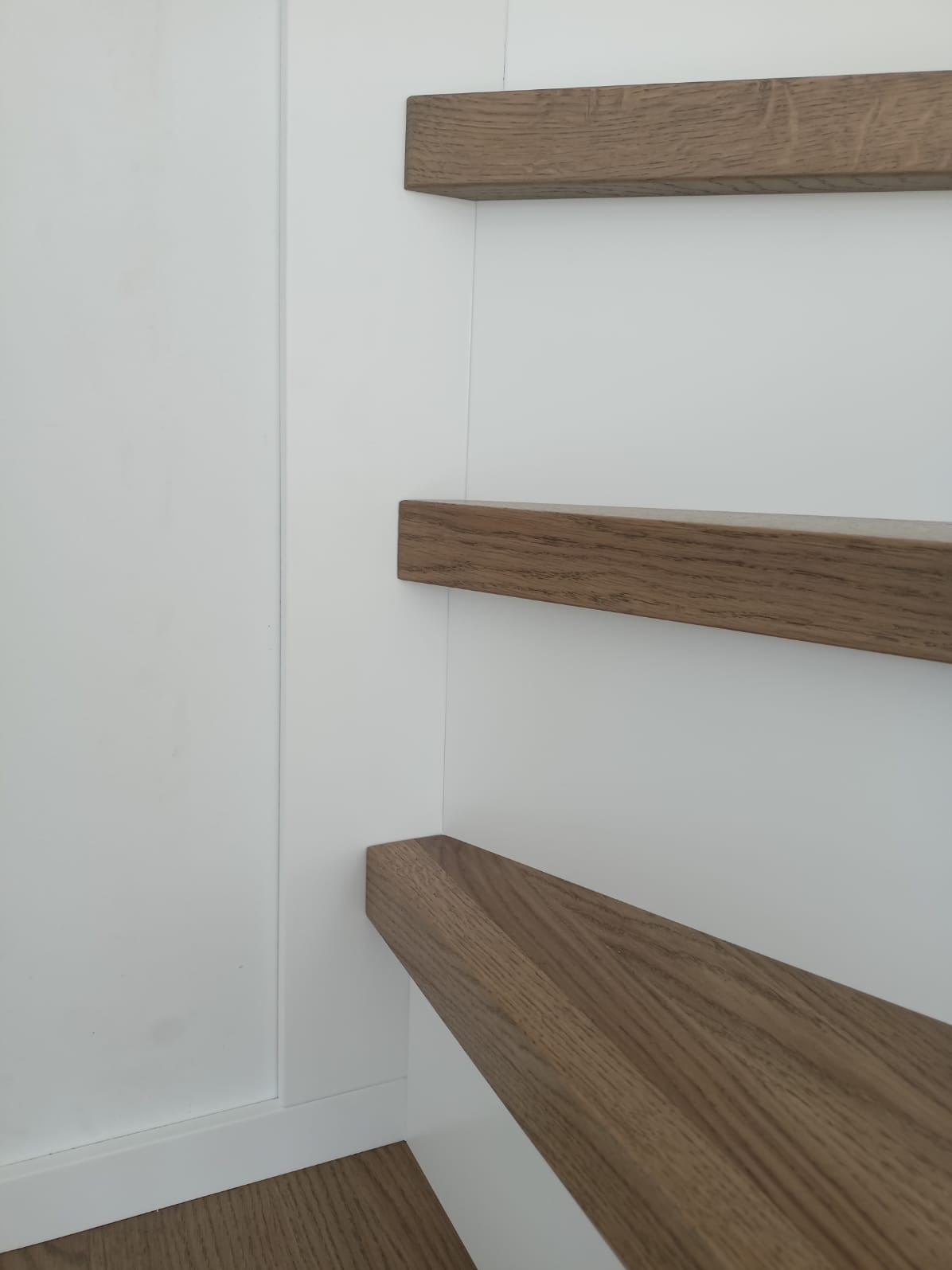 Minimalistyczne, drewniane schody wspornikowe przy białej ścianie. Stopnie z wyraźnym usłojeniem drewna, brak poręczy, nowoczesny design.