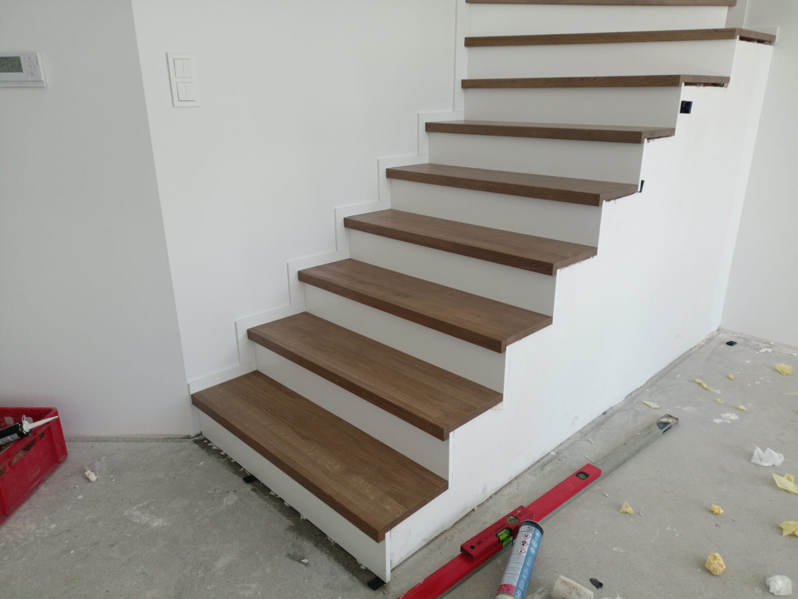 Nowoczesne schody z drewnianymi stopniami i białymi podstopnicami, widoczne narzędzia montażowe na surowej podłodze, świeżo po instalacji.