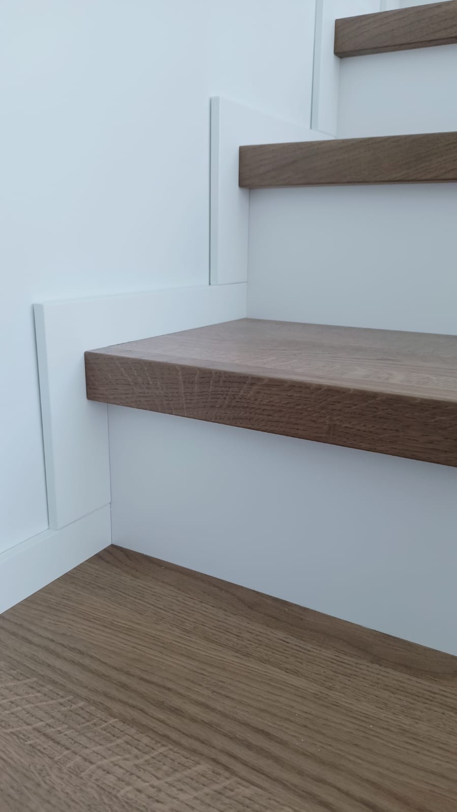 Detale drewnianych schodów z białymi panelami, widoczne stopnie i fragment podłogi, minimalistyczny design, naturalne oświetlenie.