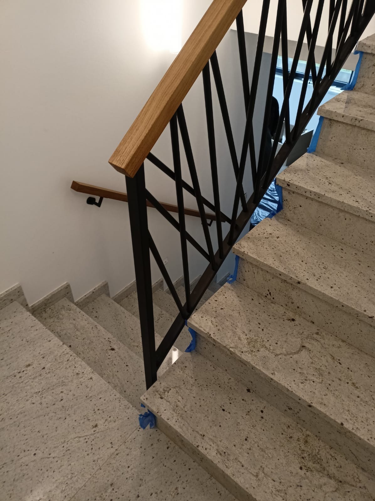 Granitowe schody z czarną, metalową balustradą i drewnianym poręczą, zabezpieczone niebieską taśmą malarską podczas montażu. Widok z góry na dół.