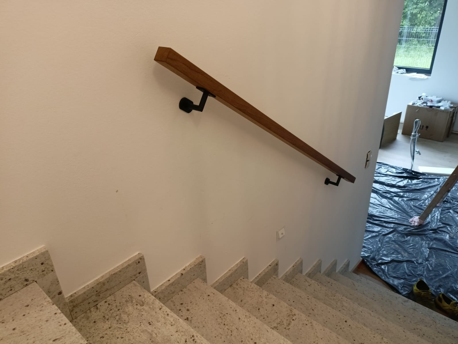 Granitowe schody z drewnianą poręczą na ścianie. Widok z góry, lekko z boku. W tle zabezpieczona folią podłoga.