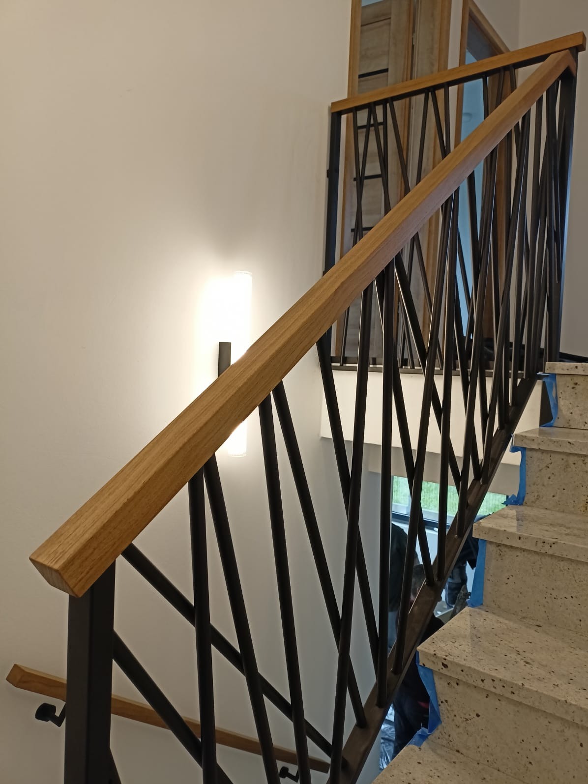 Nowoczesne schody z drewnianą poręczą i metalową balustradą o geometrycznym wzorze, oświetlone lampą ścienną, prowadzące na wyższe piętro. Detal wykończenia.