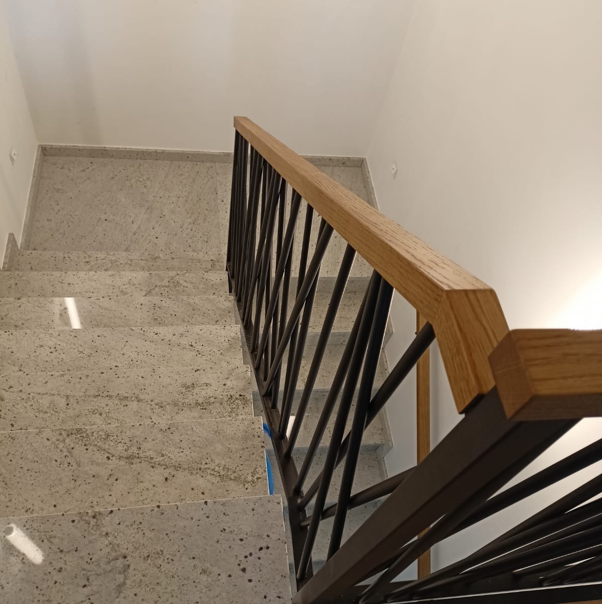 Nowoczesne schody z metalową balustradą o geometrycznym wzorze i drewnianym poręczą, widziane z góry. Stopnie wyłożone jasnym kamieniem.