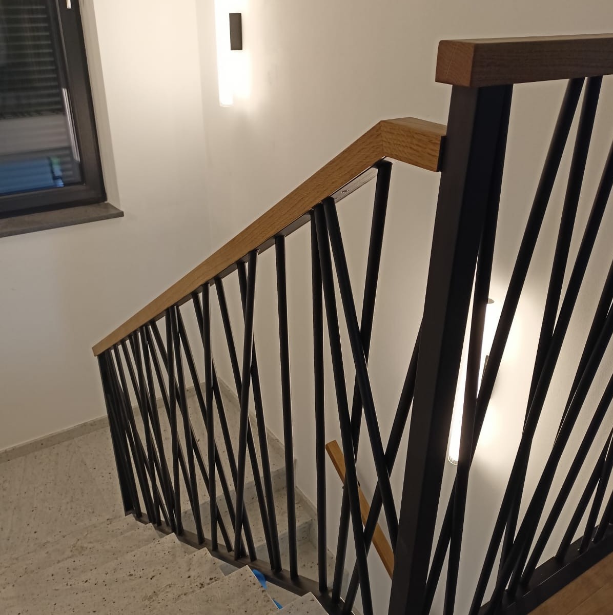 Nowoczesne schody z czarną metalową balustradą o geometrycznym wzorze i drewnianym pochwytem, oświetlone przez minimalistyczne lampy ścienne.