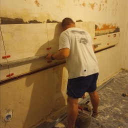 G-M-BUD - Mężczyzna w trakcie układania płytek na nierównej ścianie, widoczne krzyżyki dystansowe, poziomica i wiadro z zaprawą.