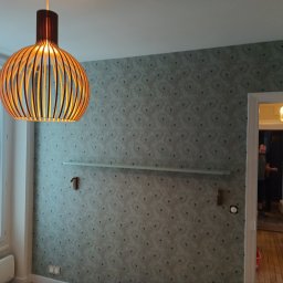 G-M-BUD - Pokój po remoncie z wzorzystą tapetą w odcieniach szarości i błękitu, drewniana lampa wisząca, okno z ozdobną balustradą i widoczne przygotowania do montażu półki.