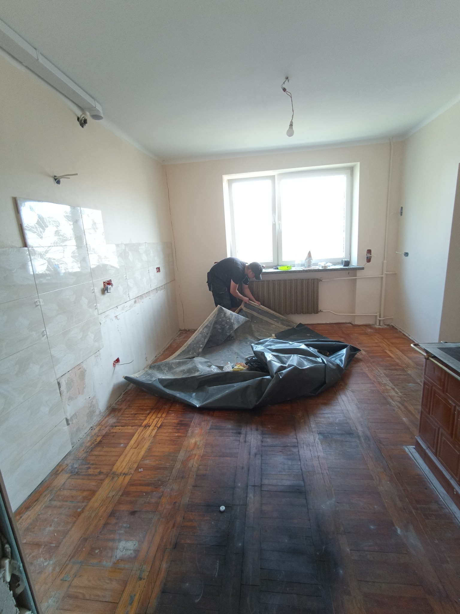 Przygotowanie pomieszczenia do prac remontowych: mężczyzna rozkłada folię ochronną na podłodze z parkietu, ściany częściowo pokryte płytkami, widoczne puszki elektryczne.