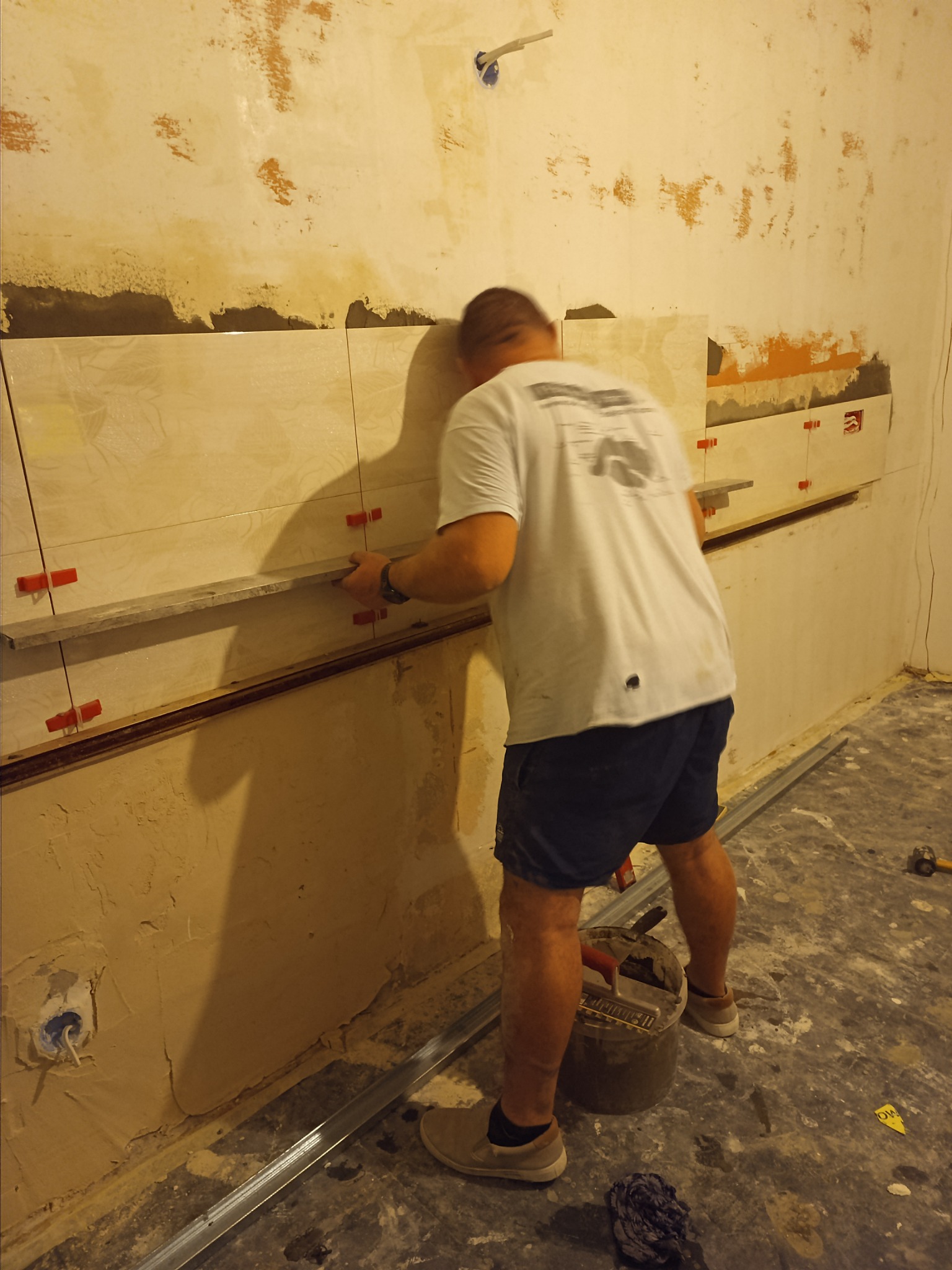 Mężczyzna w trakcie układania płytek na nierównej ścianie, widoczne krzyżyki dystansowe, poziomica i wiadro z zaprawą.