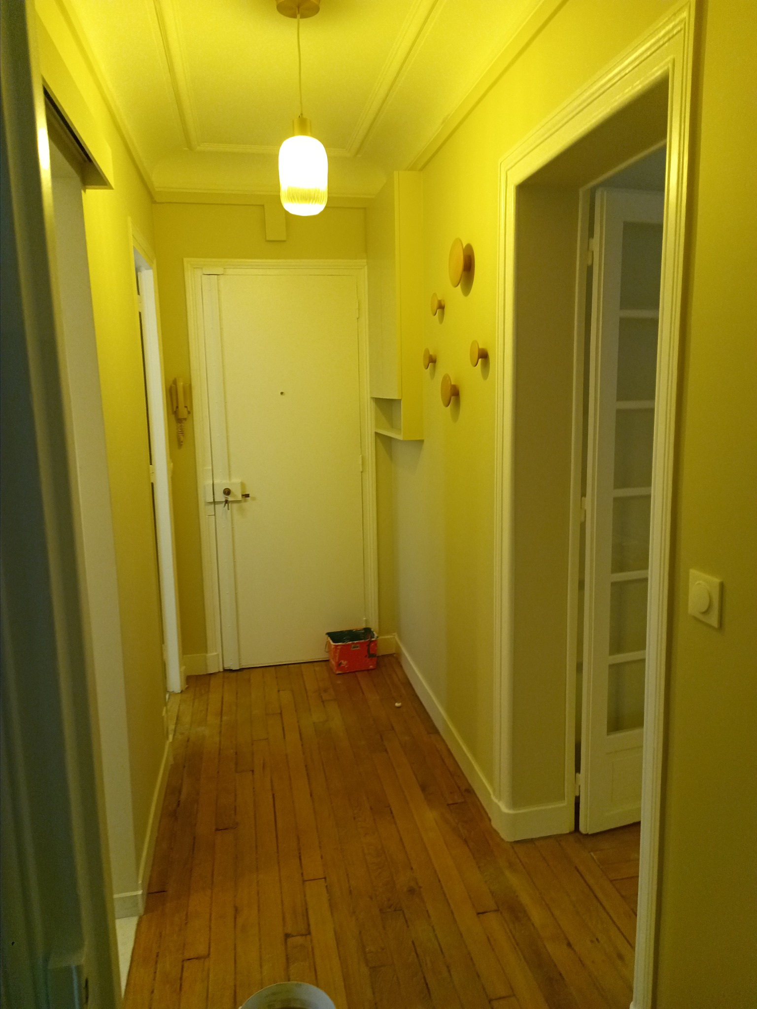 Wnętrze przedpokoju z drewnianą podłogą i żółtymi ścianami, widoczne drzwi wejściowe, drzwi wewnętrzne, wieszaki na ścianie i lampa sufitowa.