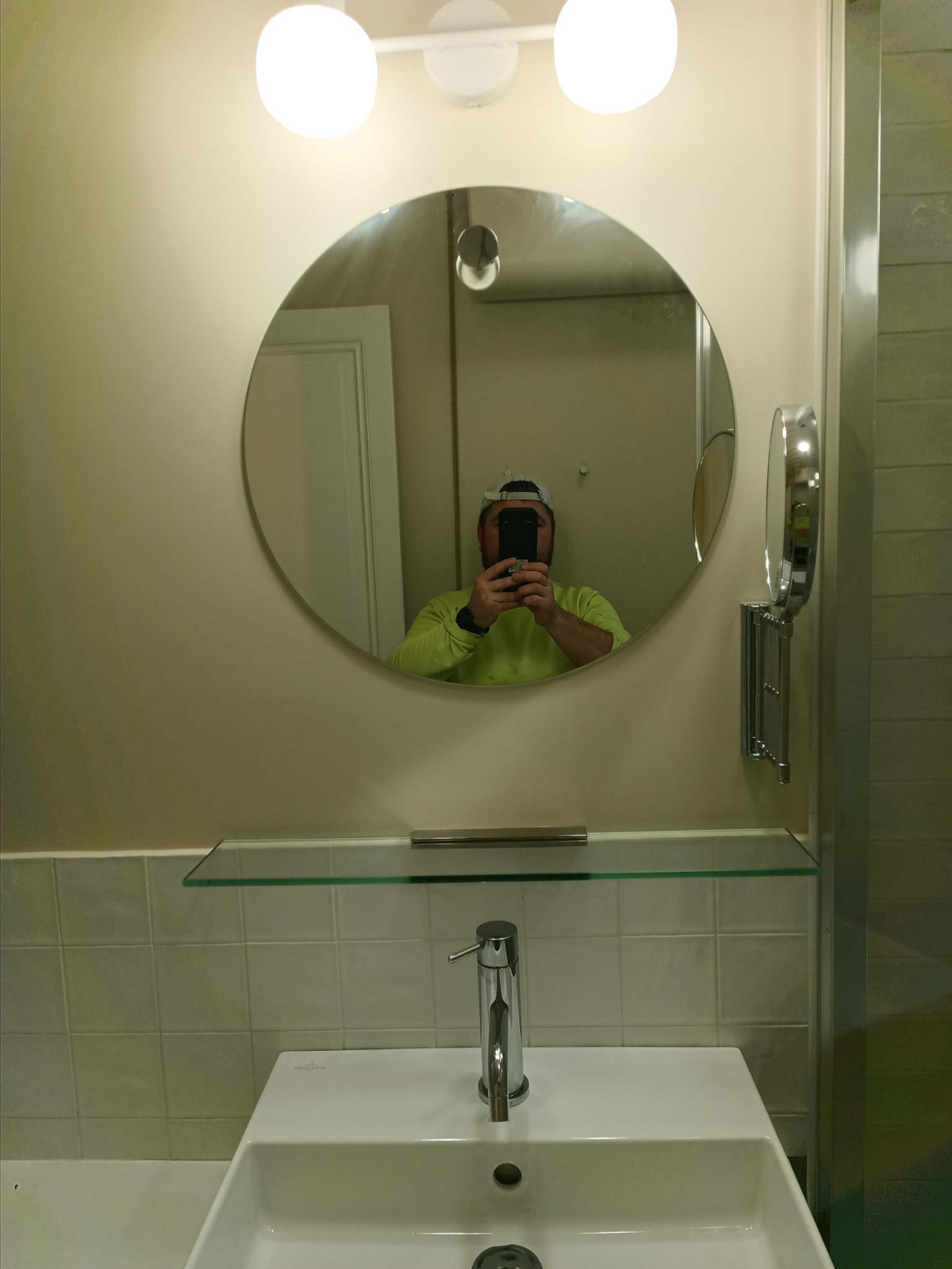 Nowoczesna umywalka z baterią, okrągłe lustro z podświetleniem i regulowane lusterko kosmetyczne w odnowionej łazience, widoczne odbicie osoby robiącej zdjęcie.