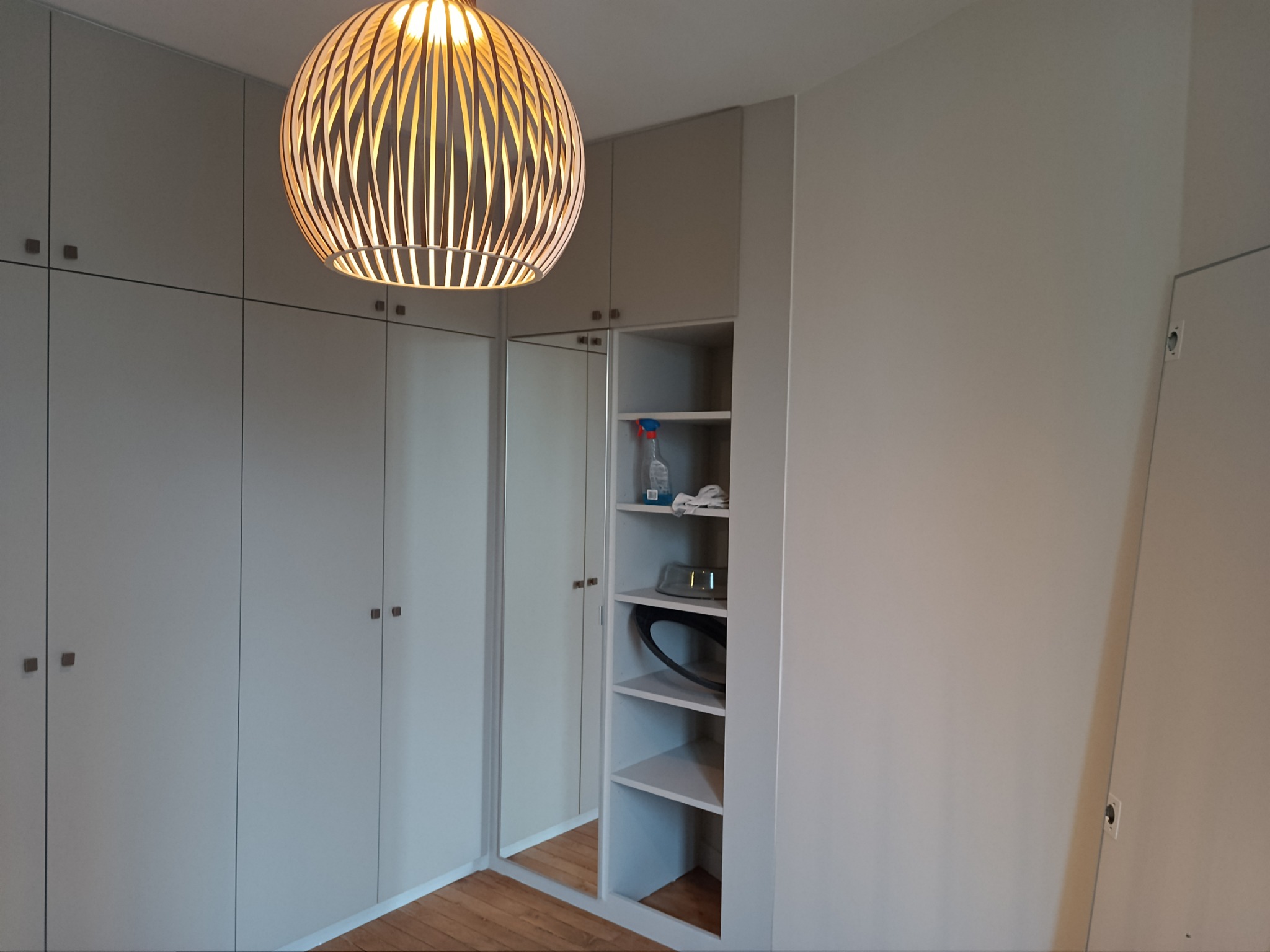 Wnętrze pokoju z zabudową szaf w jasnych odcieniach szarości, drewniana podłoga i dekoracyjna lampa wisząca z ażurowym kloszem.