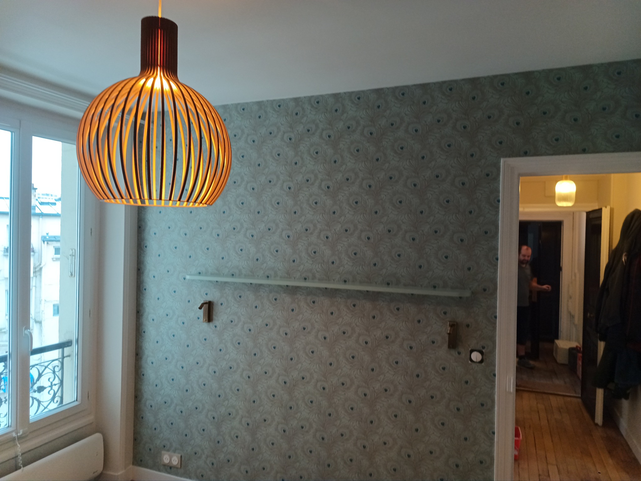 Pokój po remoncie z wzorzystą tapetą w odcieniach szarości i błękitu, drewniana lampa wisząca, okno z ozdobną balustradą i widoczne przygotowania do montażu półki.